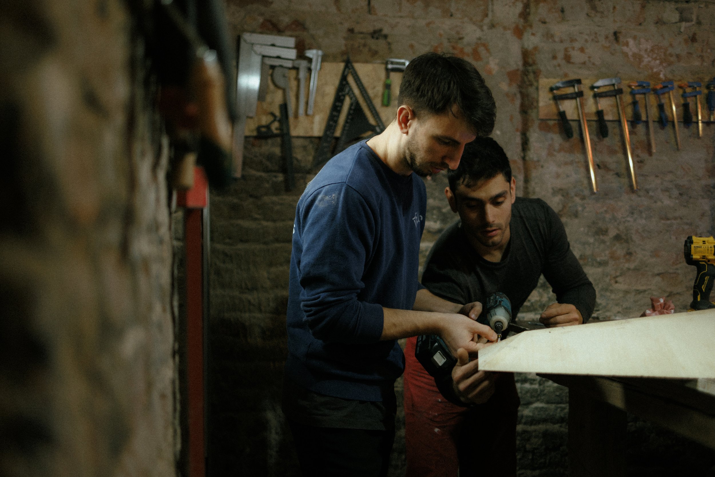 Two men working with a cordless drill on a wooden board in a workshop, surrounded by tools hanging on the wall.