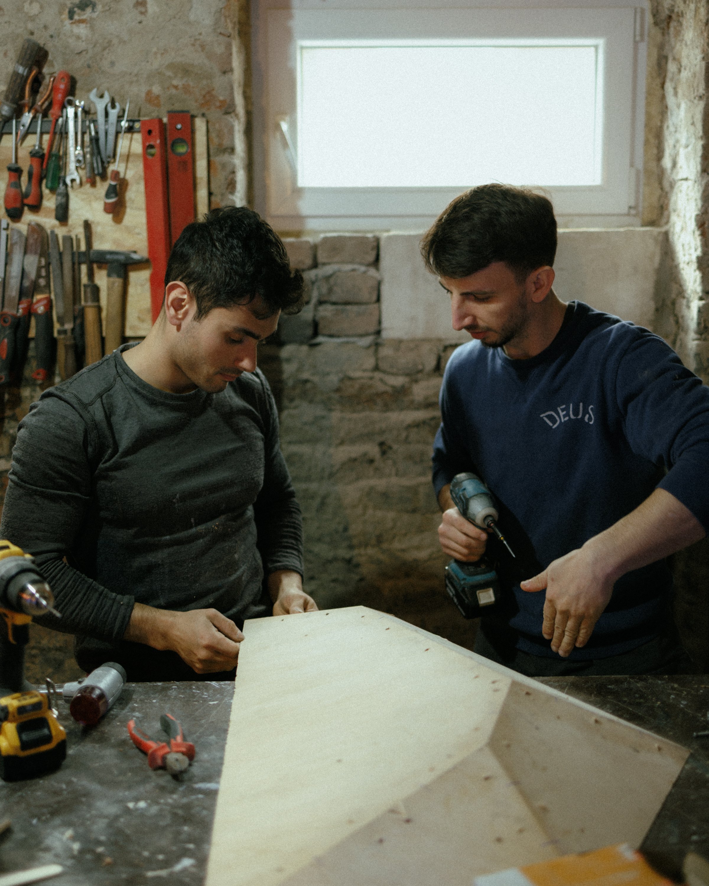 Two men working on a woodworking project in a workshop. They are using a drill and other tools. Various tools hang on the wall in the background.