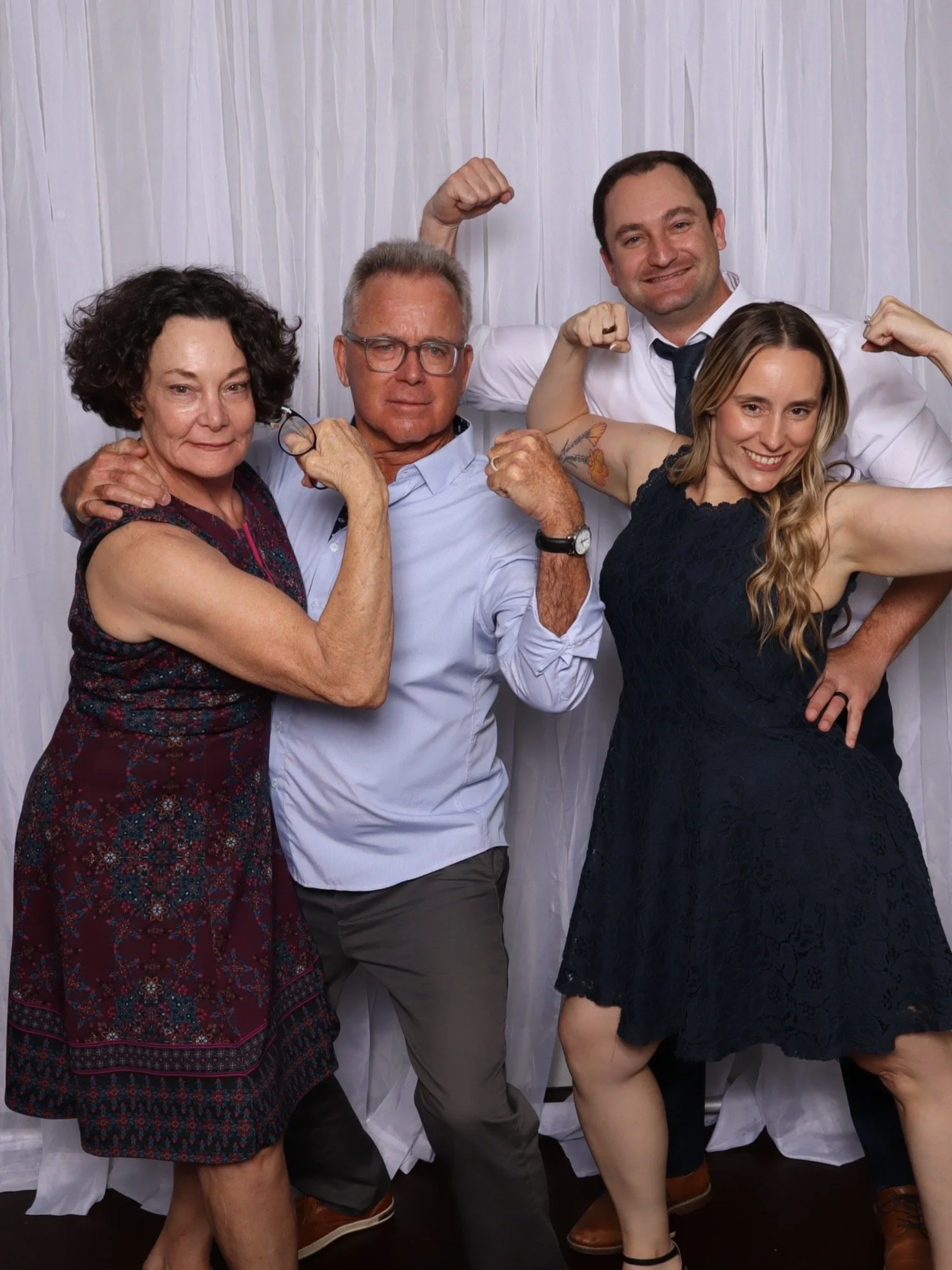 Group of four people posing together, some with flexed muscles, in front of a white curtain background.