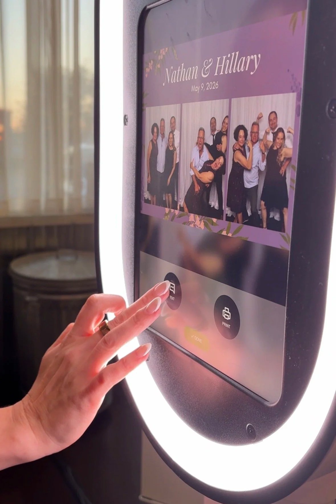 A person using a touchscreen photo kiosk showing a wedding photo collage labeled 'Nathan & Hillary' with the date May 9, 2026, and buttons for 'Text' and 'Print'.