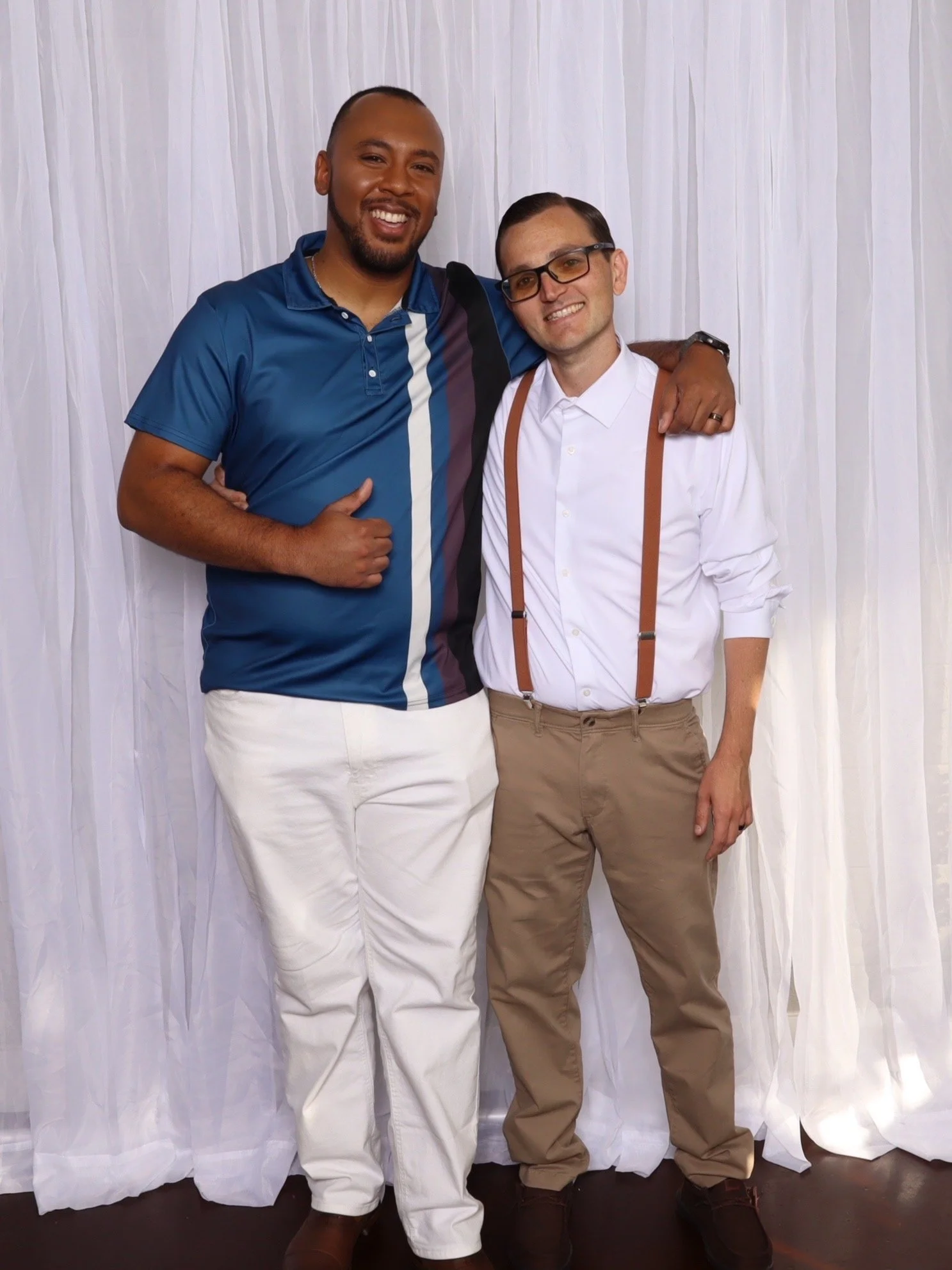 Two men smiling and hugging each other in front of a white curtain background. One man is wearing a blue shirt and white pants, and the other is wearing a white shirt with suspenders and khakis.