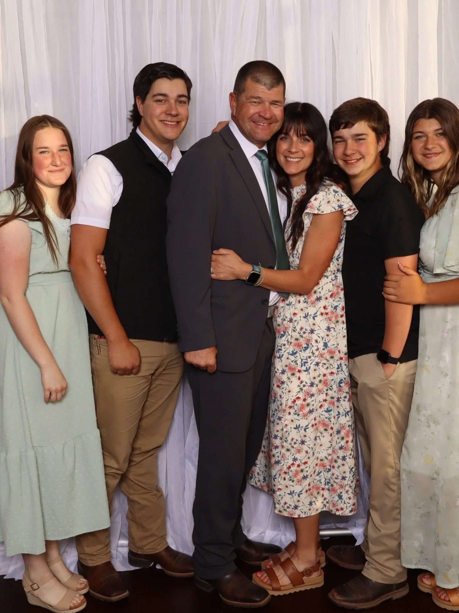 A family of six people standing close together, smiling, in front of a white curtain backdrop, dressed in semi-formal and casual clothing.