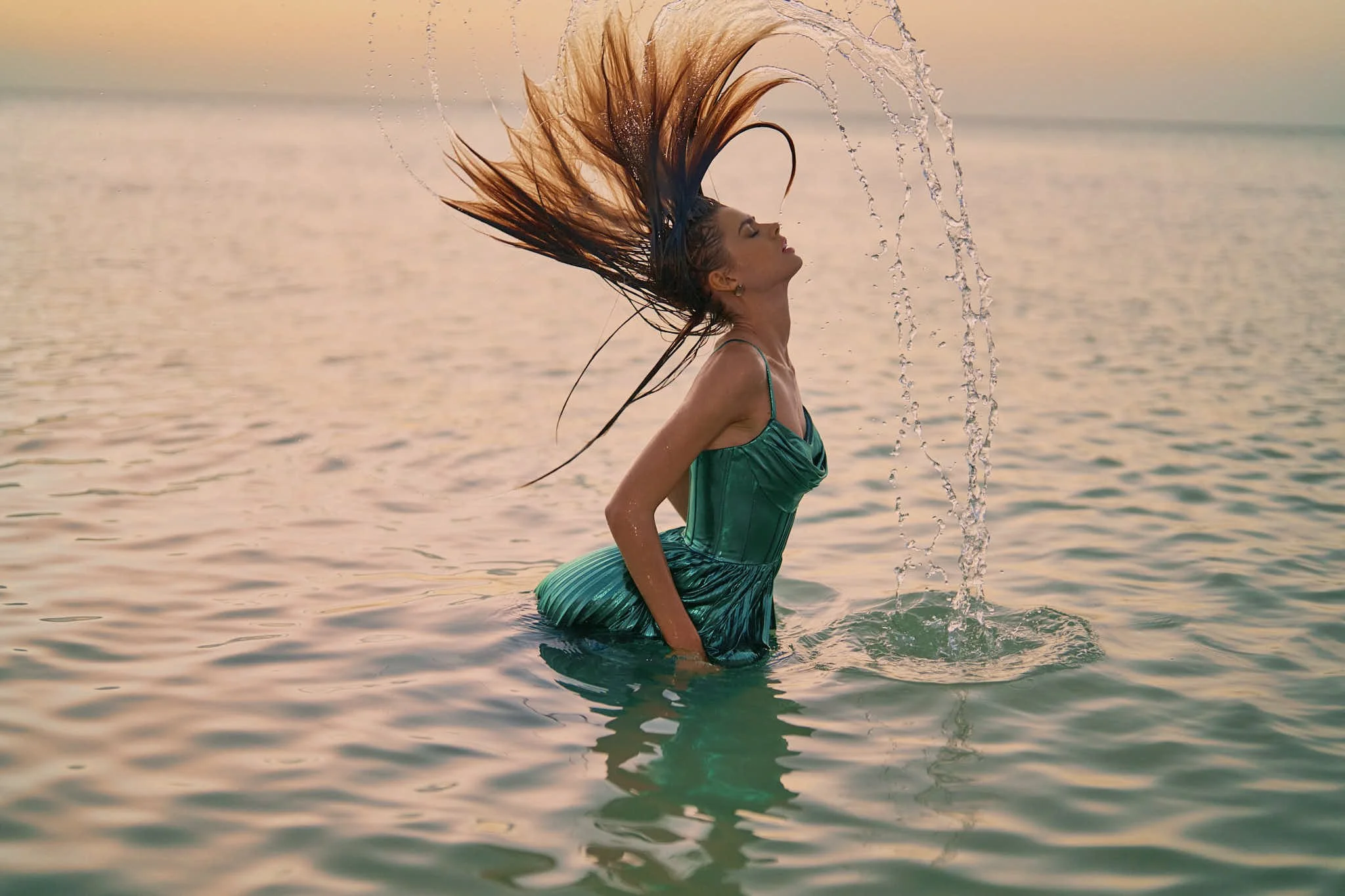 A woman in a teal dress stands in water, flipping her wet hair back, creating an arc of water droplets, during sunset on a calm body of water.