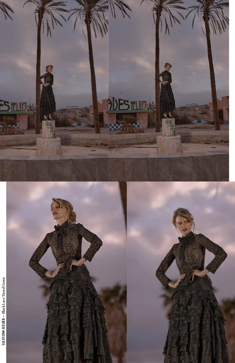 A woman posing outdoors at dusk in a black lace long dress with ruffles, standing on a concrete pedestal against palm trees and cloudy sky.