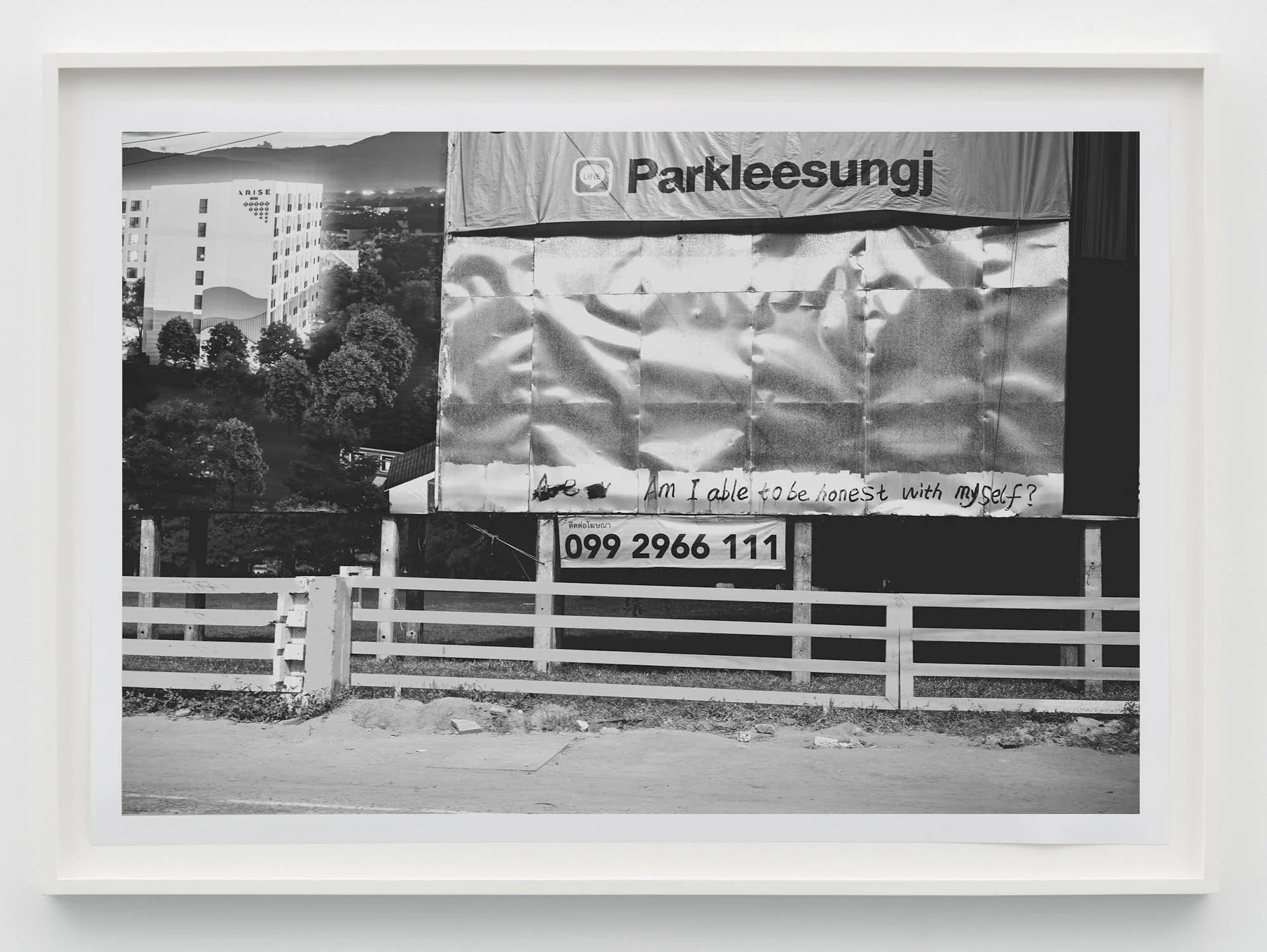 A black and white photo of a billboard with a background of trees and buildings, with a message written in marker on the billboard that says, 'Am I able to be honest with myself?' and a phone number below.