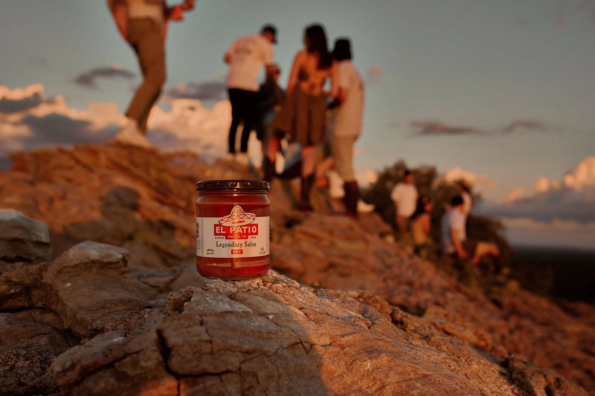 Jar of El Patio salsa placed on rocky terrain with a group of people standing and sitting on a rocky hillside in the background during sunset.