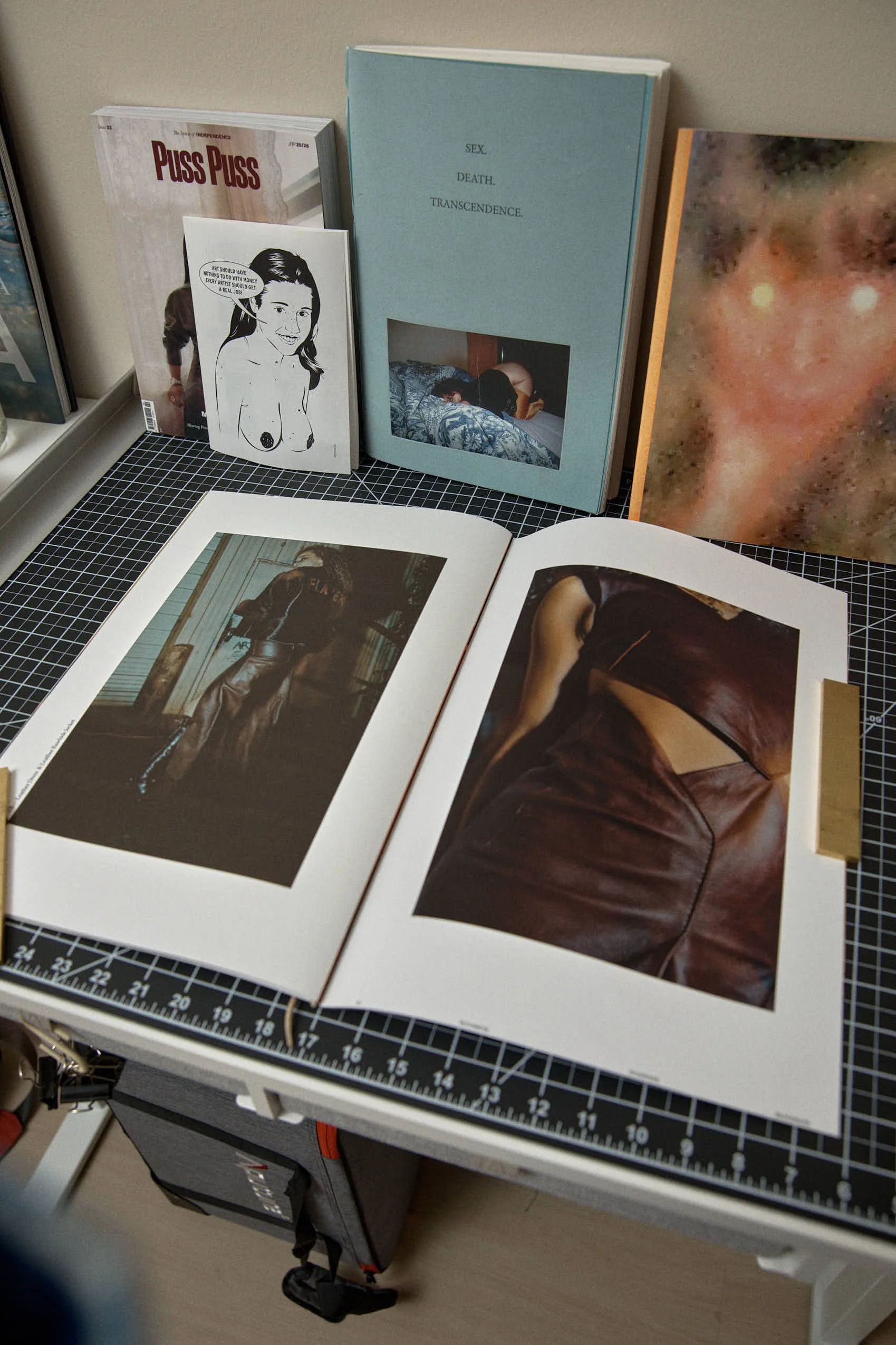 A display of art books and magazines, including photographs of women wearing leather clothing, on a black grid cutting mat.