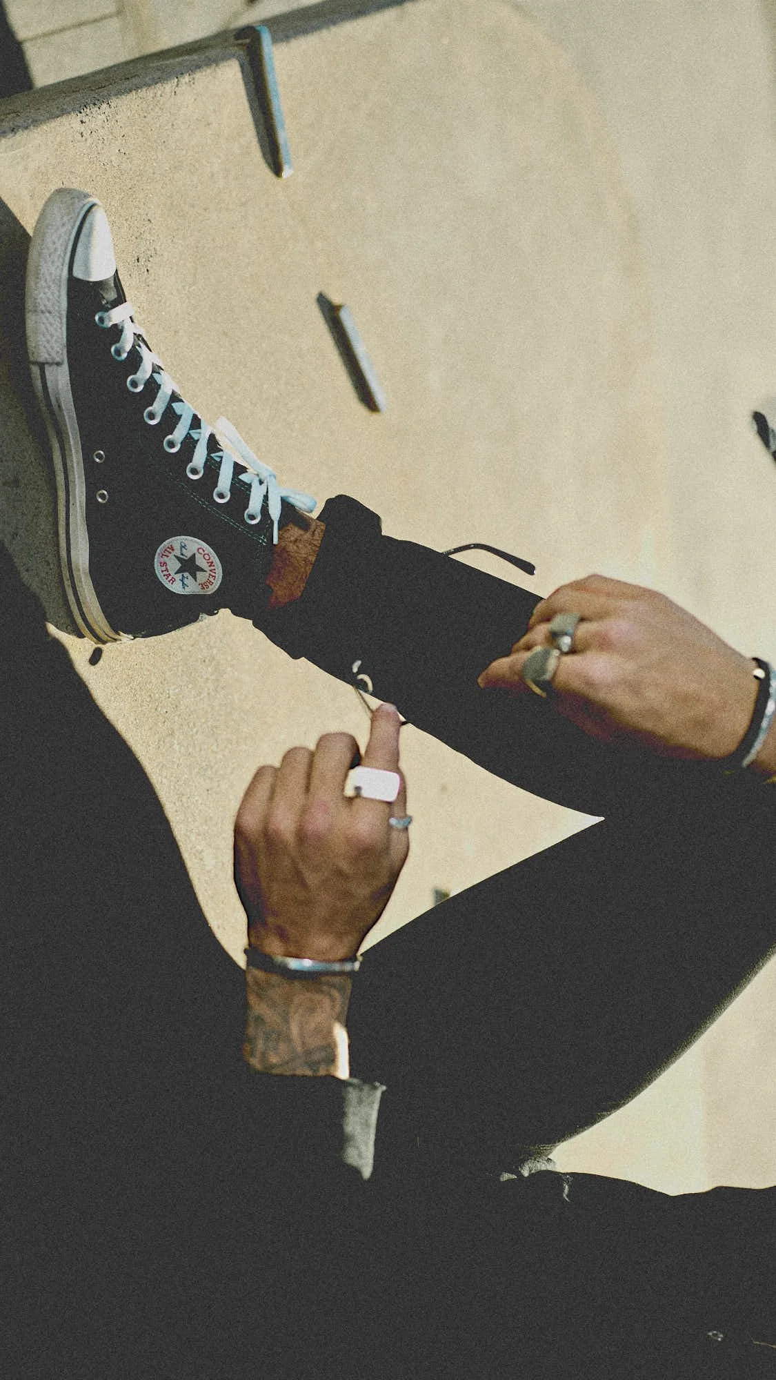 Person sitting on the ground, wearing a Converse Chuck Taylor All Star sneaker, adjusting the laces.