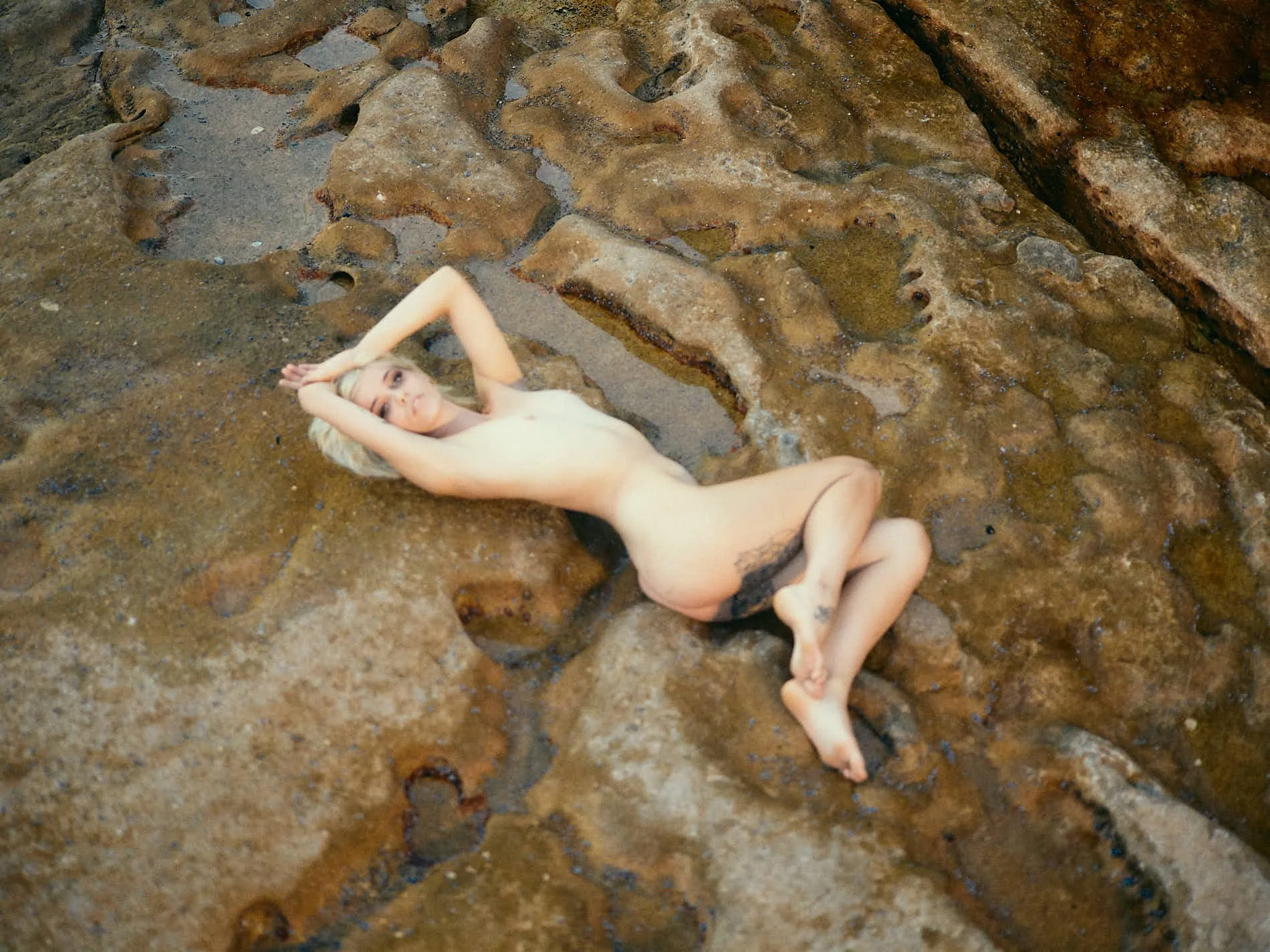 Nude woman lying on a rocky surface with arms above her head, in an outdoor setting.