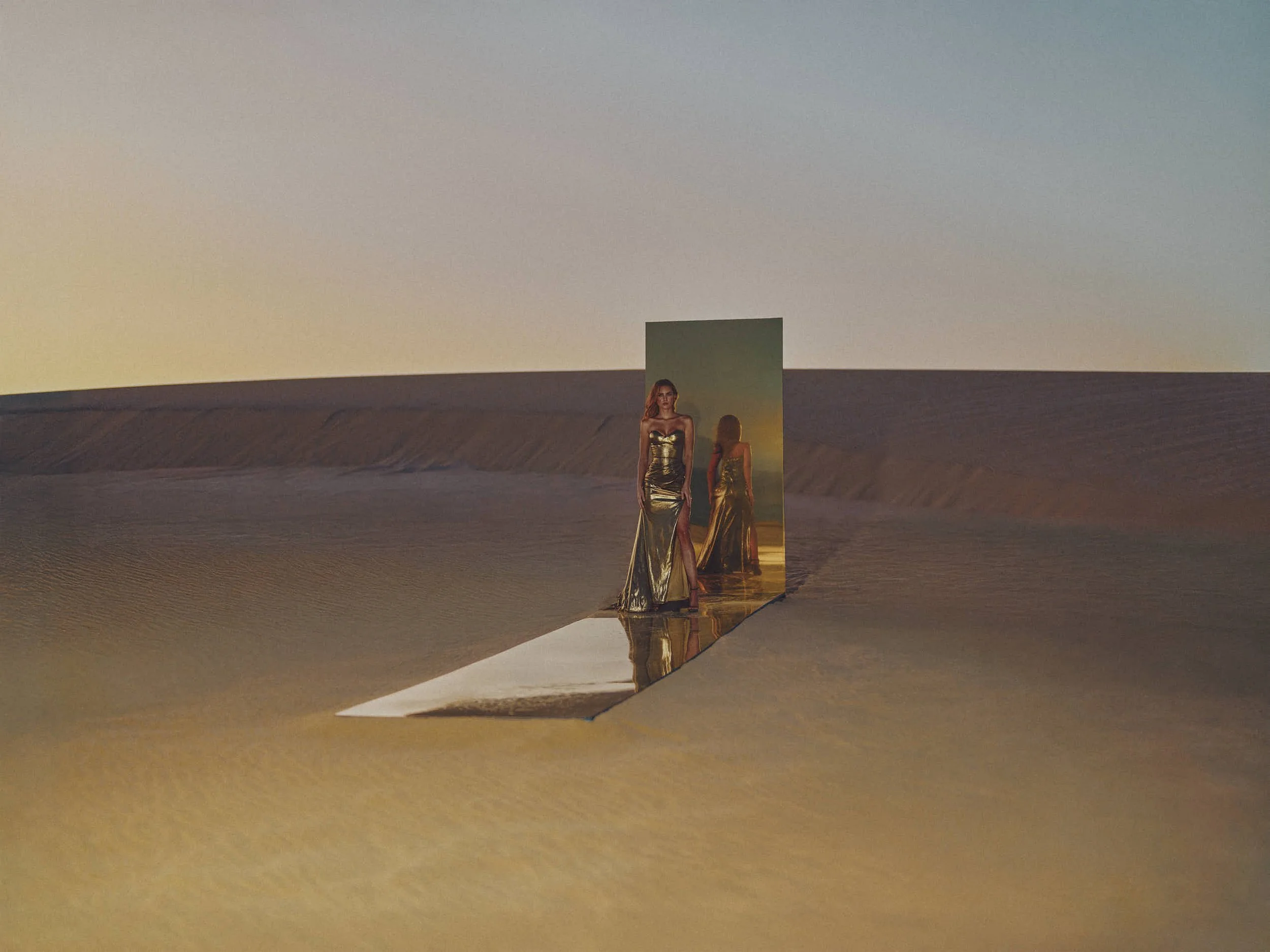 A mirror aligned with the sandy desert landscape reflects a woman in a gold gown at sunset, creating an illusion of her standing in the desert.