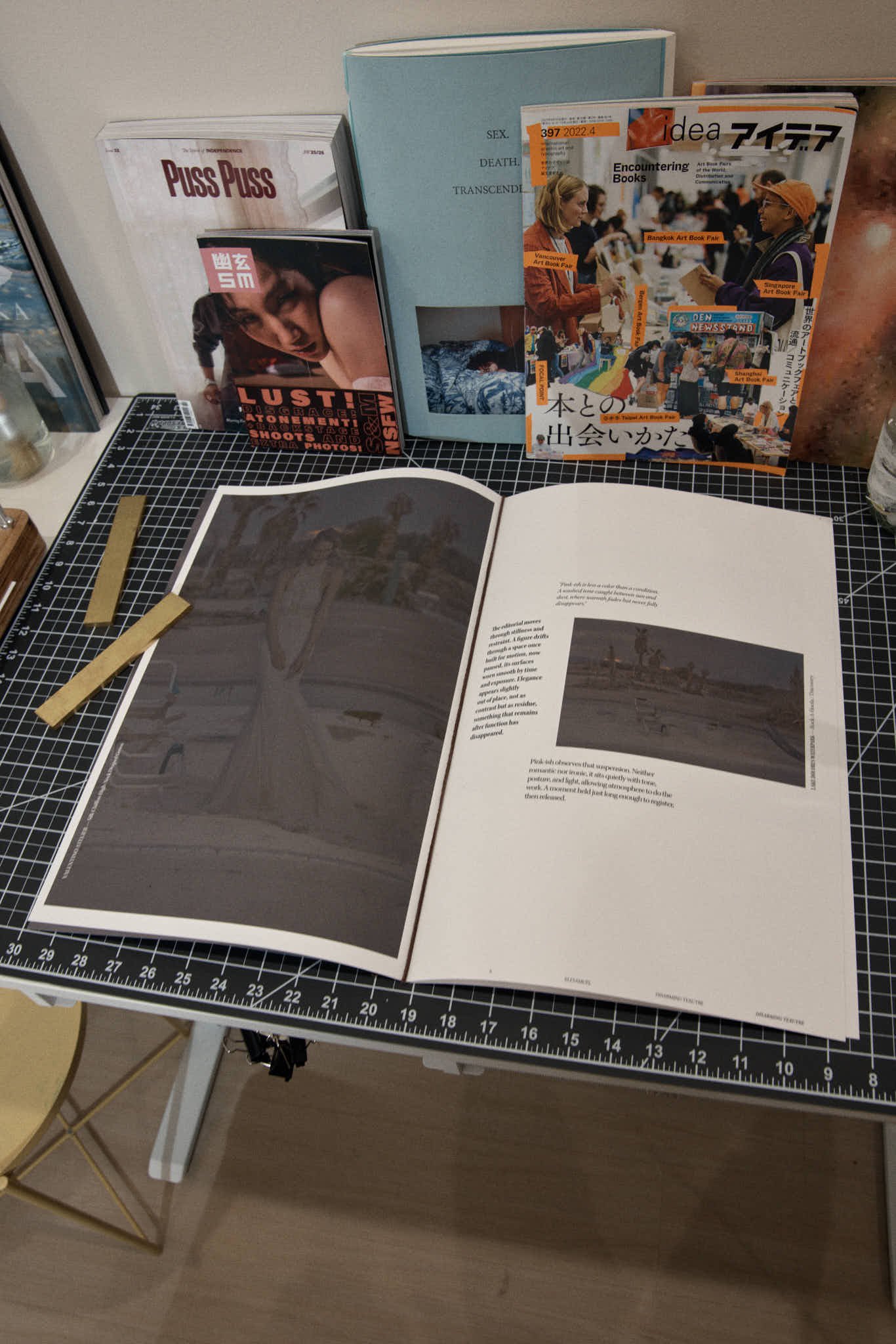 Open magazine on a black grid cutting mat with thick beige borders and black clips, showing a two-page spread with black-and-white photographs of a landscape and some text.
