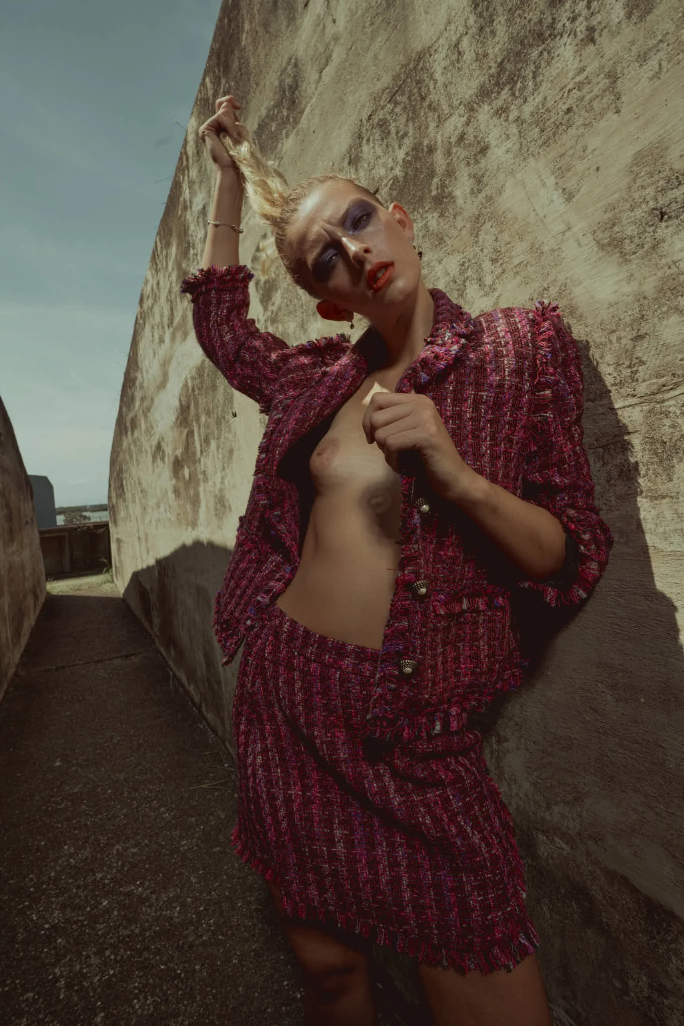 A woman in a pink tweed suit with a matching skirt, posing against a concrete wall outdoors during daytime. She has platinum blonde hair tied up and dramatic makeup with bold eyes and lipstick.