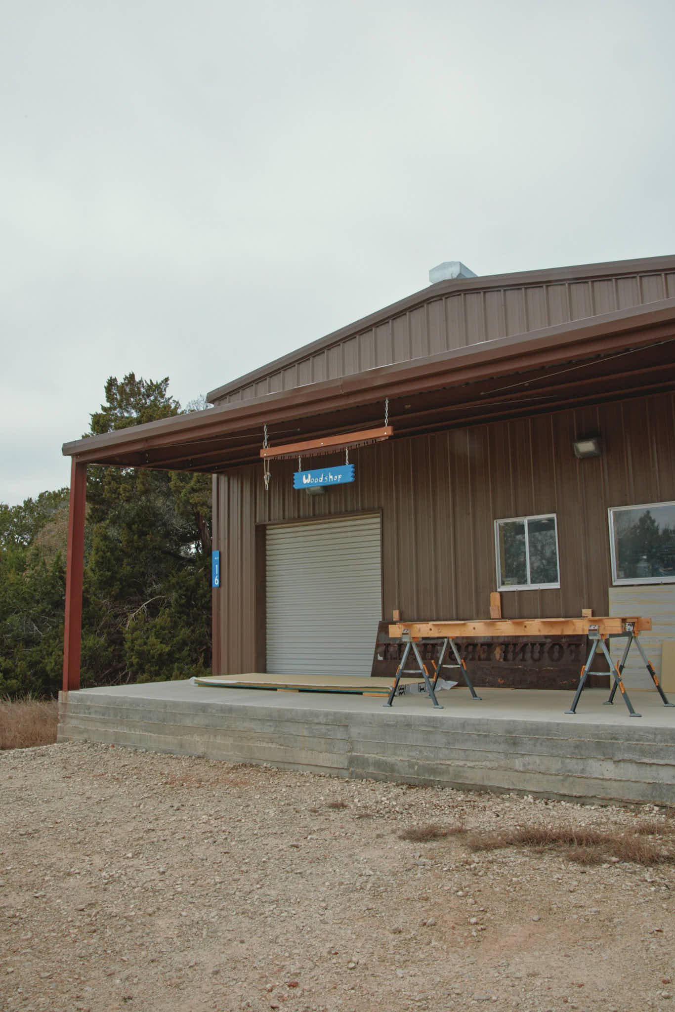 Exterior of a woodworking shop with a metal building, a partially open rolling door, and wooden workbenches on a concrete platform.