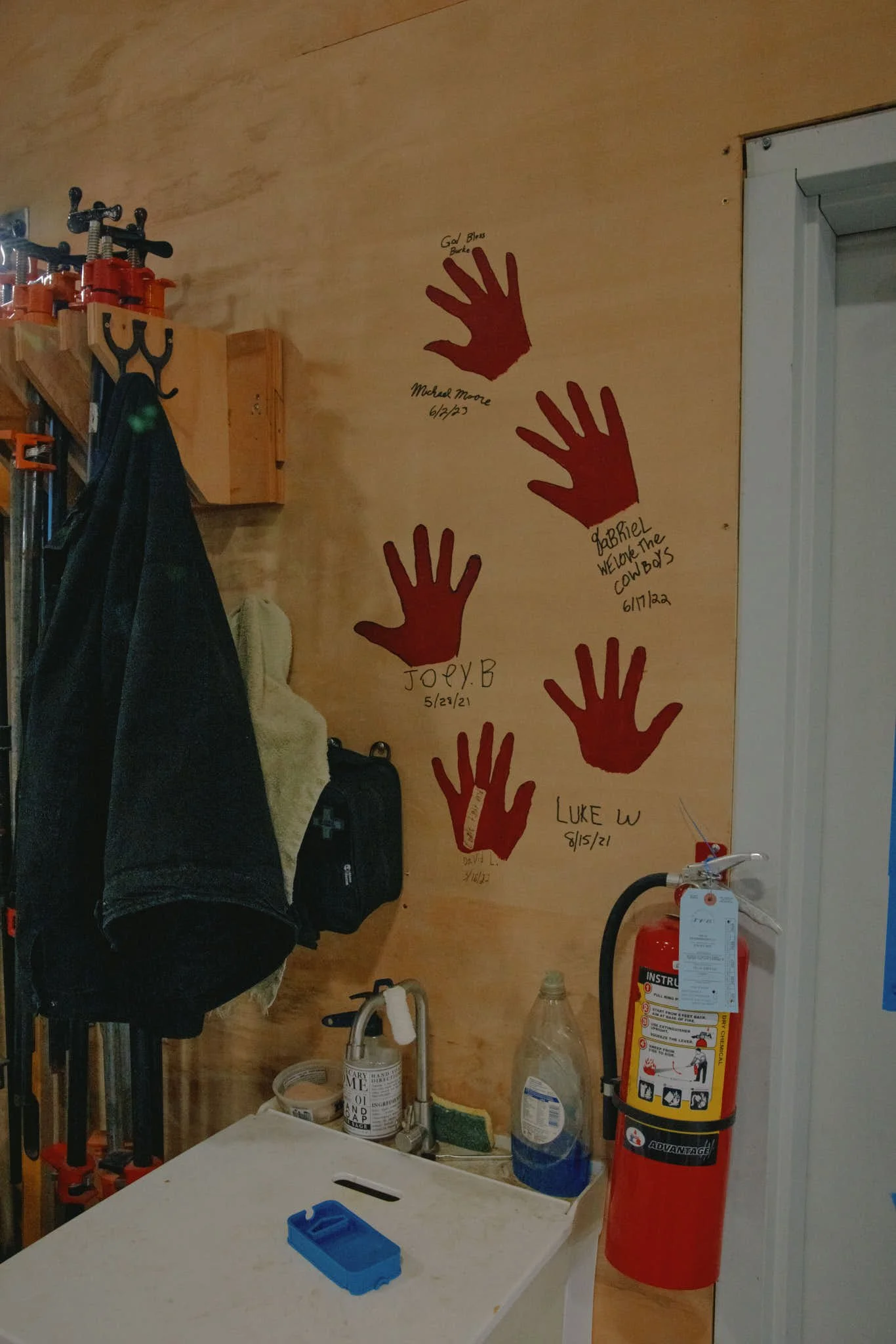 Painted red handprints on a wooden wall with signatures and dates beneath each handprint, located in a workshop or garage setting.