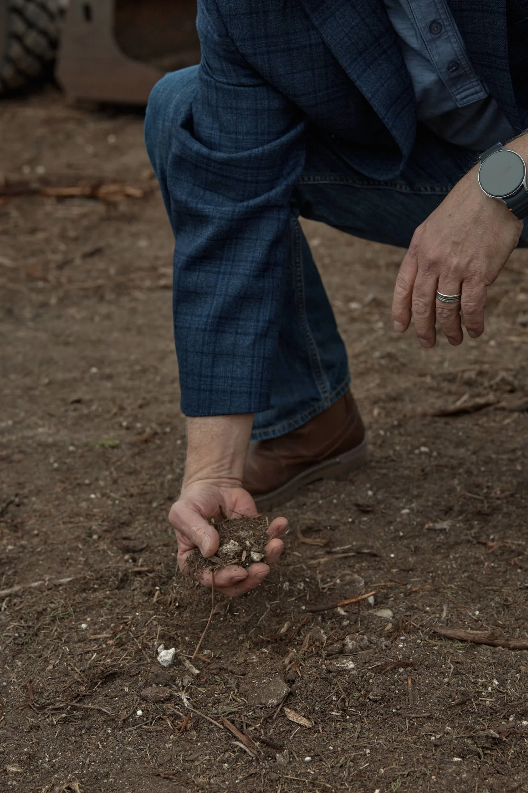 A person dressed in a blue plaid suit, with a watch on their wrist and a ring on their finger, is crouched down on the ground and holding a handful of soil.