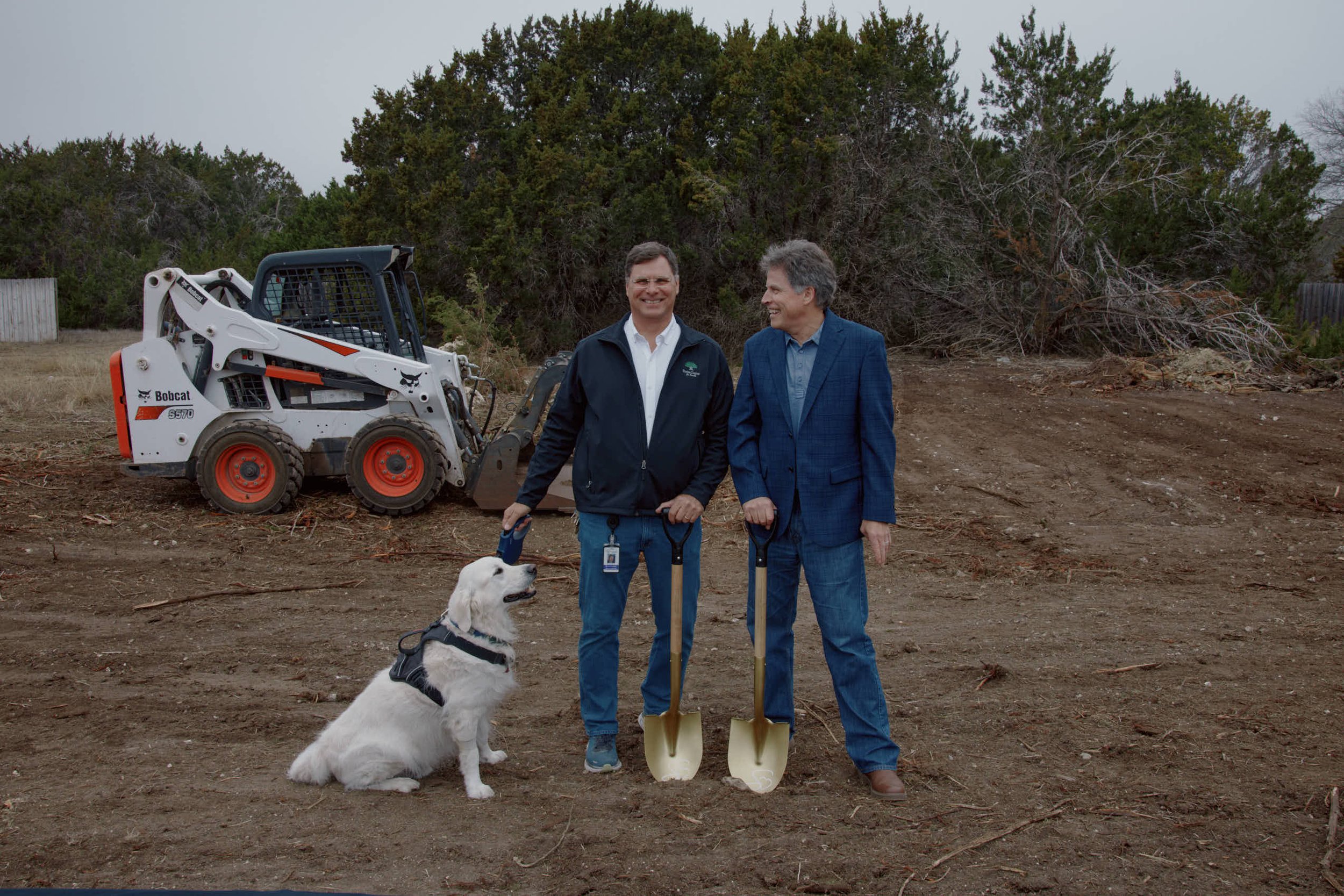 Two men standing outdoors on a dirt lot, holding shovels, with a golden retriever sitting beside them wearing a harness, and a small Bobcat skid-steer loader in the background. They are smiling and appear to be at a construction or land development site.