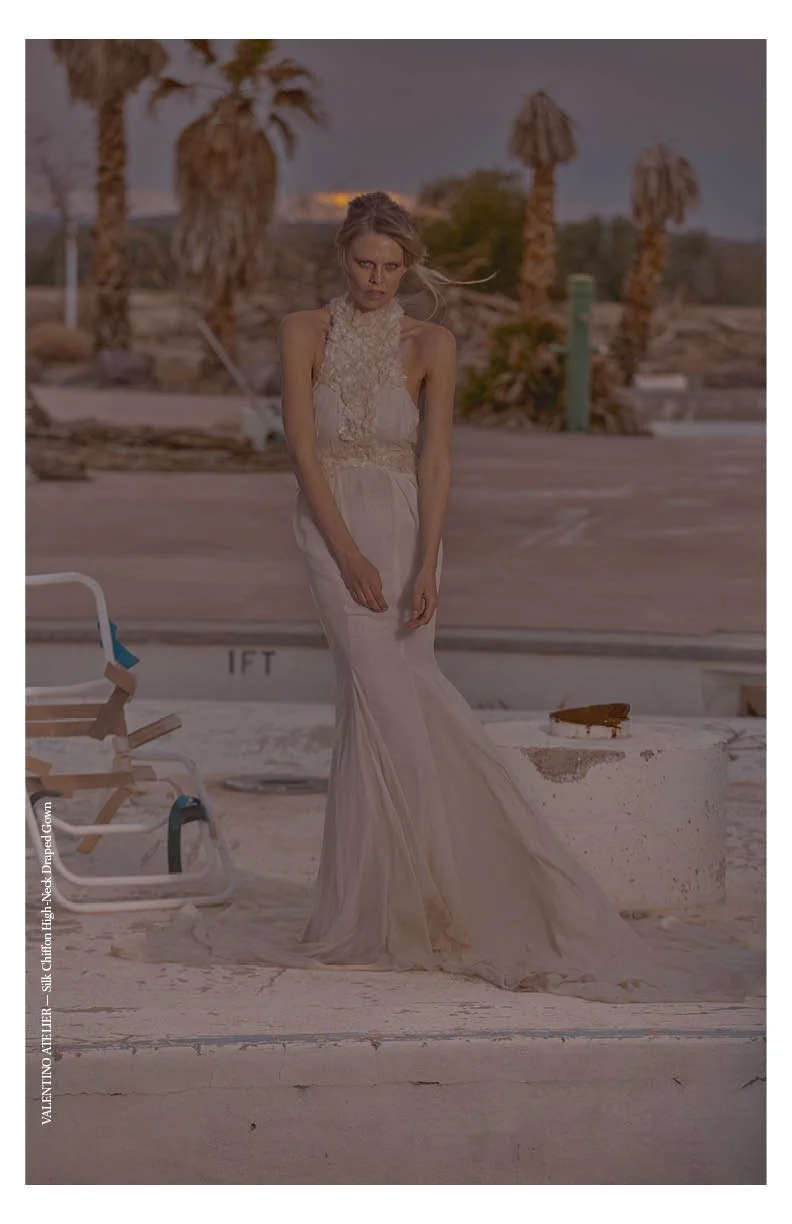 A woman in a long, elegant, beige dress standing next to a poolside in a desert-like environment with palm trees in the background, during sunset or twilight.