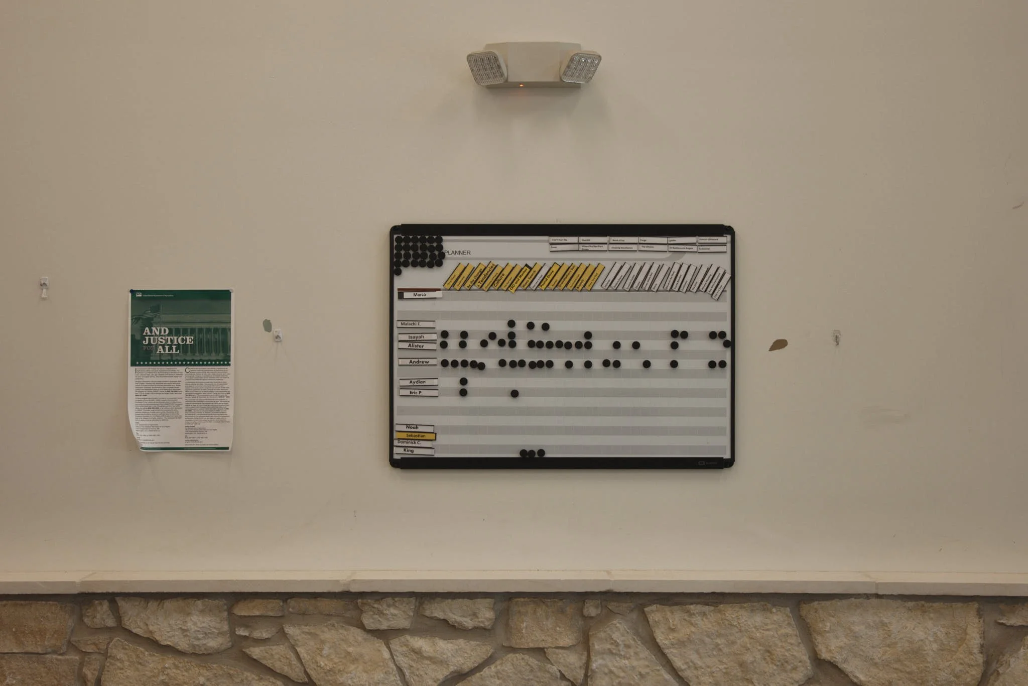 A whiteboard with black, yellow, and white magnetic blocks and markers, with some names written on it, mounted on a wall above a stone ledge, and a light fixture above it.