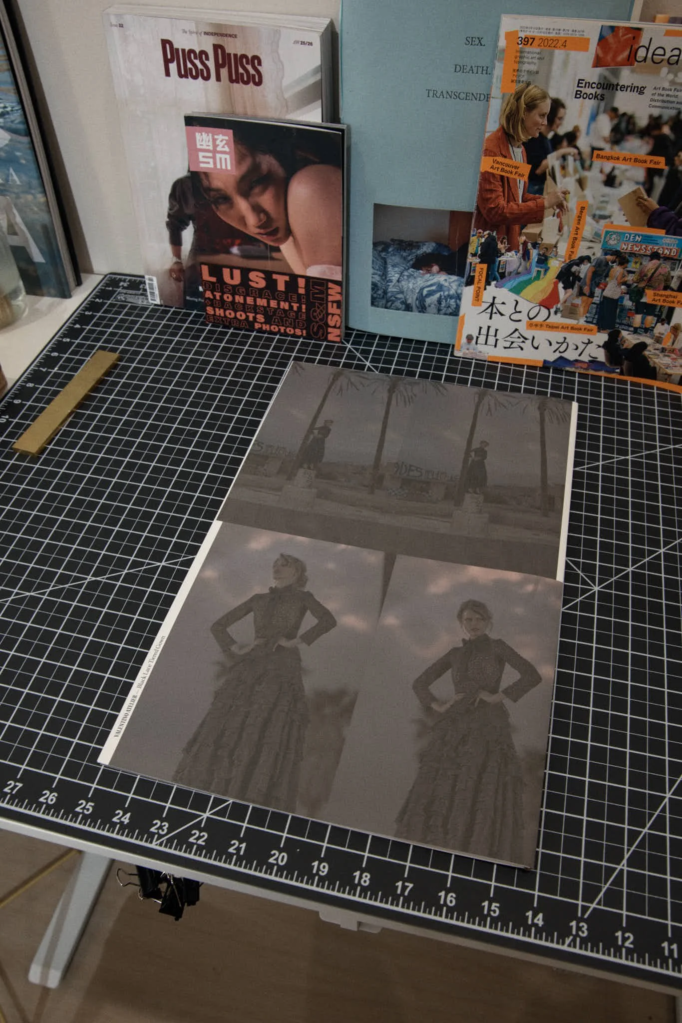 A worktable with black grid cutting mat, displaying a sheet of photo prints of a woman in a dress in various poses, with books and magazines in the background, including a magazine titled 'Puss Puss' and a book with Japanese writing and images from an art book fair.