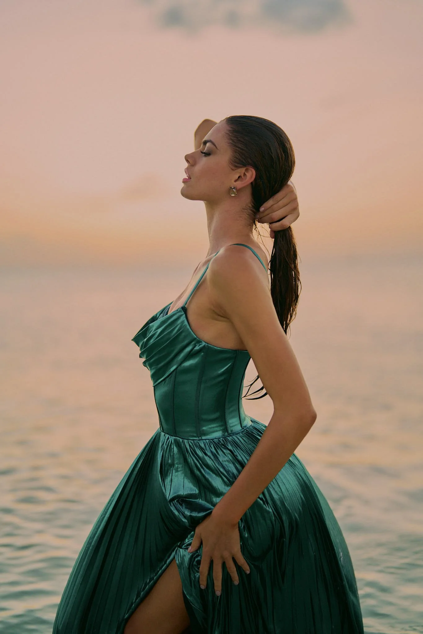 A woman in a green dress stands by the water at sunset, with her eyes closed and one hand behind her head, holding her wet hair.