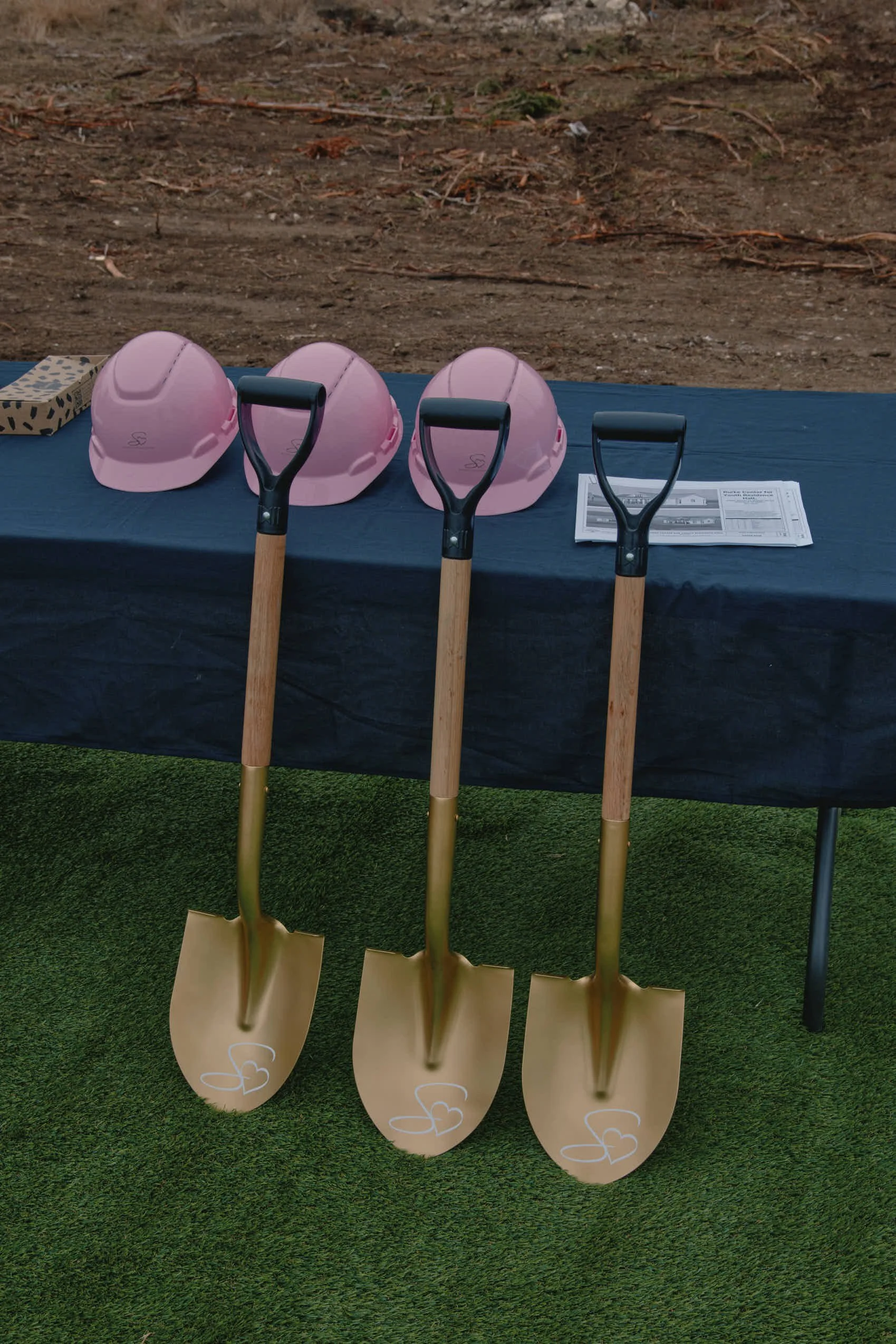 Three pink construction helmets and three gold shovels with black handles, set up at a presentation table outdoors.