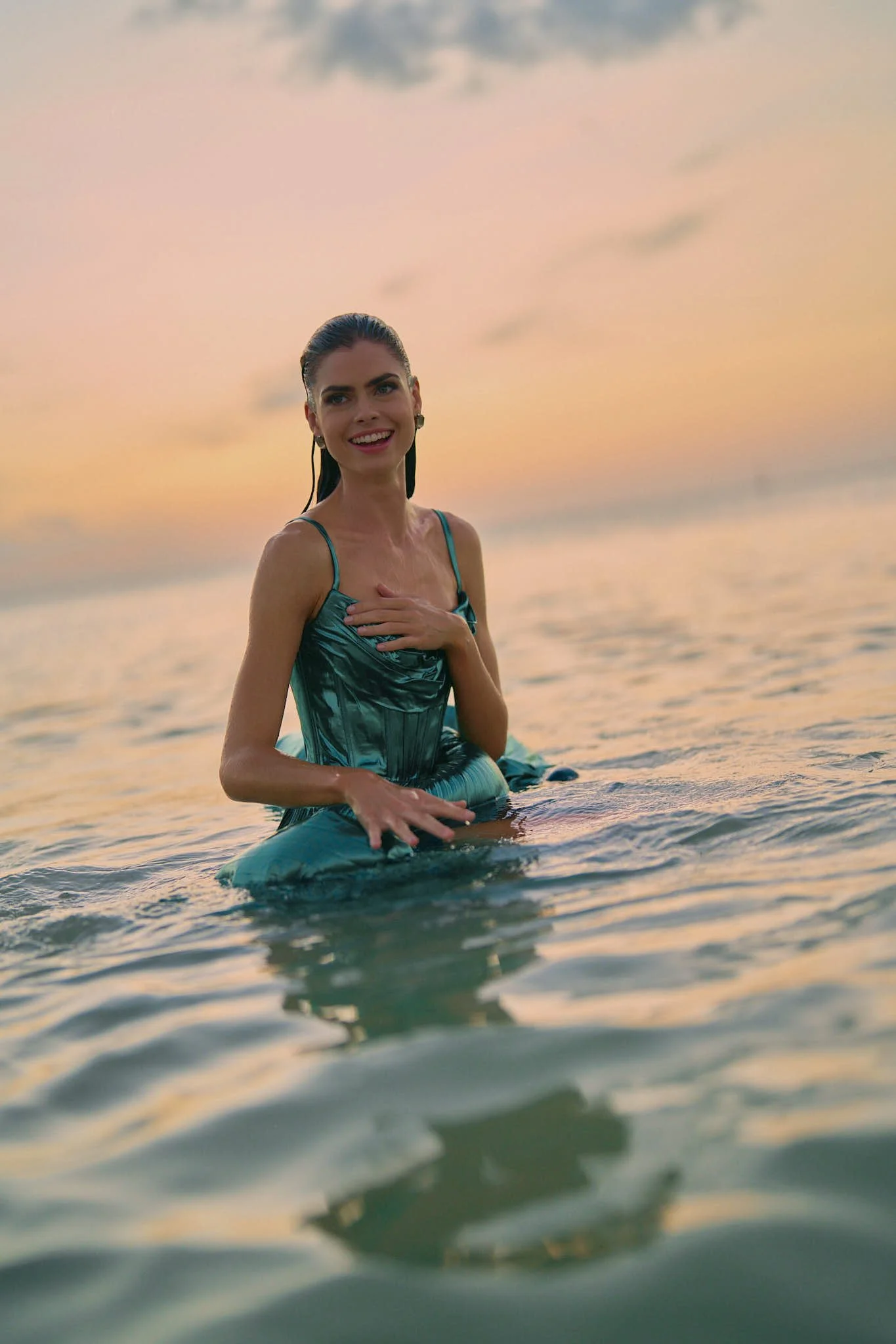 A woman in a blue dress kneeling in water at sunset, smiling and holding her chest.