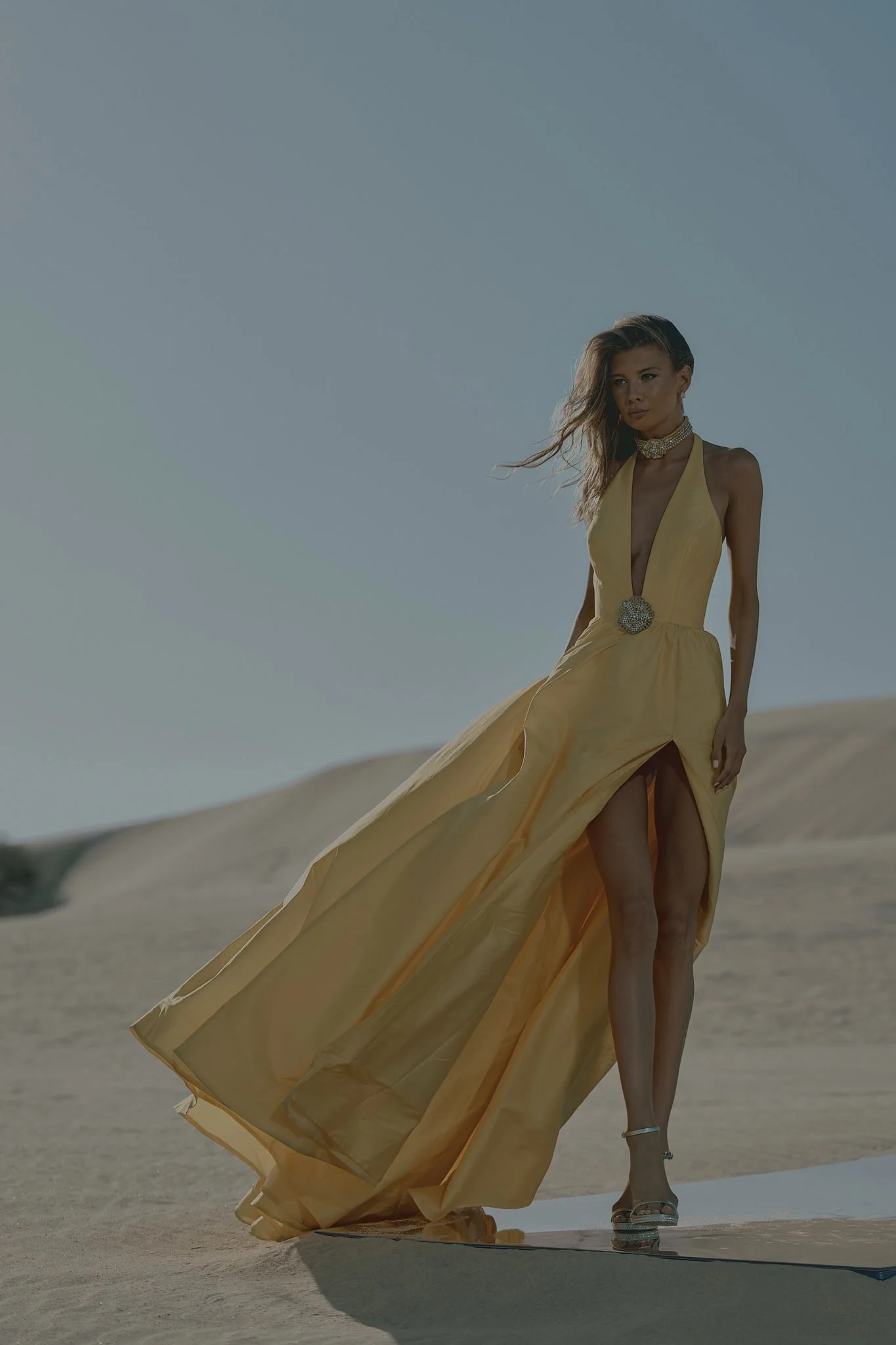 A woman walking on sand dunes wearing a yellow evening gown with high slit and high heels, with wind blowing her hair.