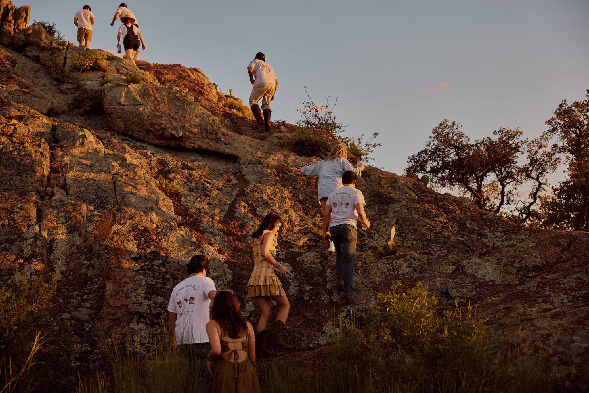 People climbing a rocky hill during sunset, some wearing casual clothes, surrounded by trees and bushes.
