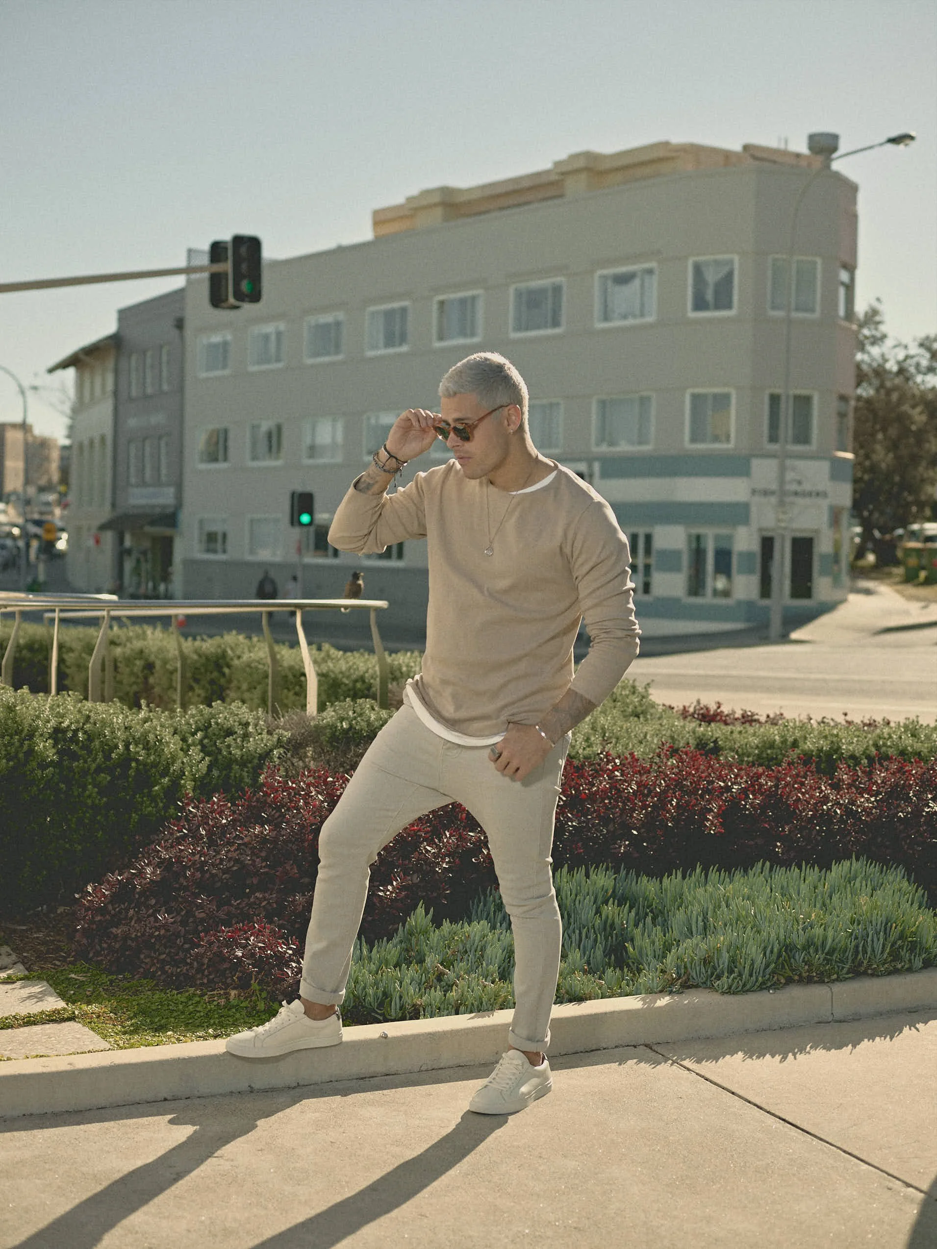 A young man with platinum blonde hair, wearing sunglasses, a beige sweatshirt, light-colored jeans, and white sneakers, standing on a sidewalk, adjusting his sunglasses with one hand and resting the other on his pocket, in an urban setting with a building and traffic light in the background during daytime.
