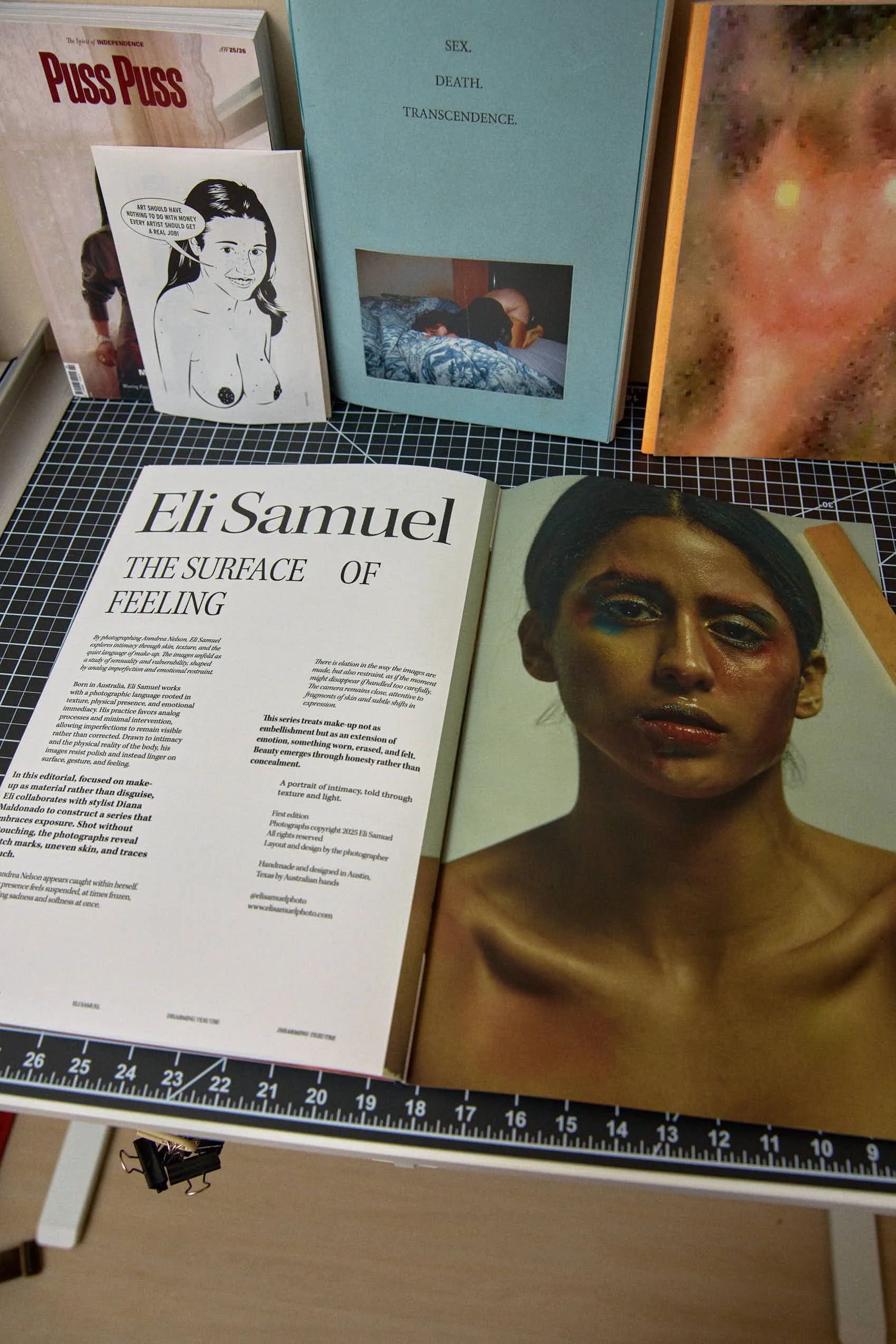 An open magazine featuring an art book with a portrait of a woman with makeup. Behind the magazine are several books and posters, including titles related to sex, death, transcendence, and political topics.