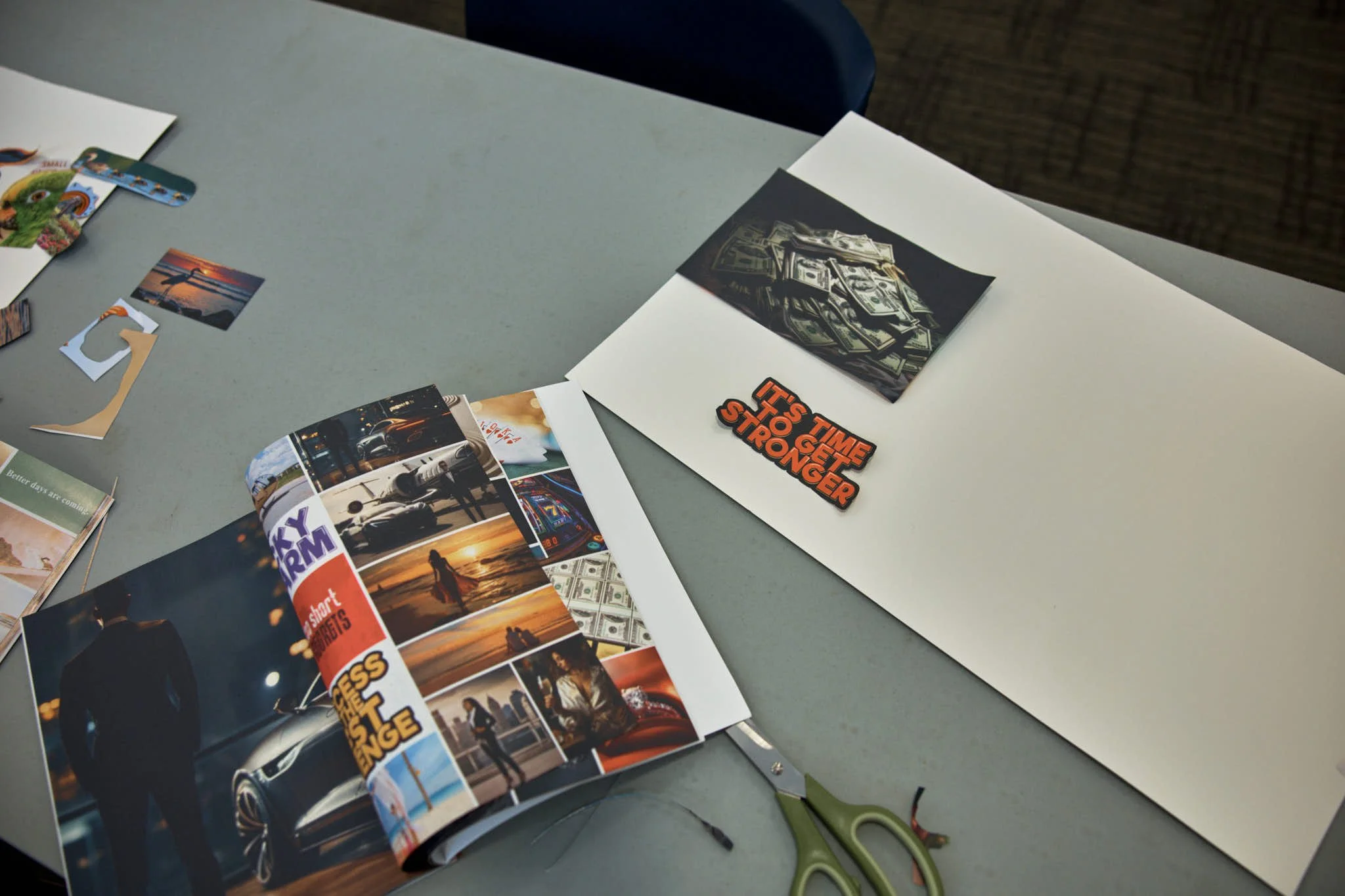 Table with various photos and images, including a stack of magazines, a photo of cash, a sticker with bold orange and black text saying 'It's Time to Get Stronger', small cutouts, and scissors.