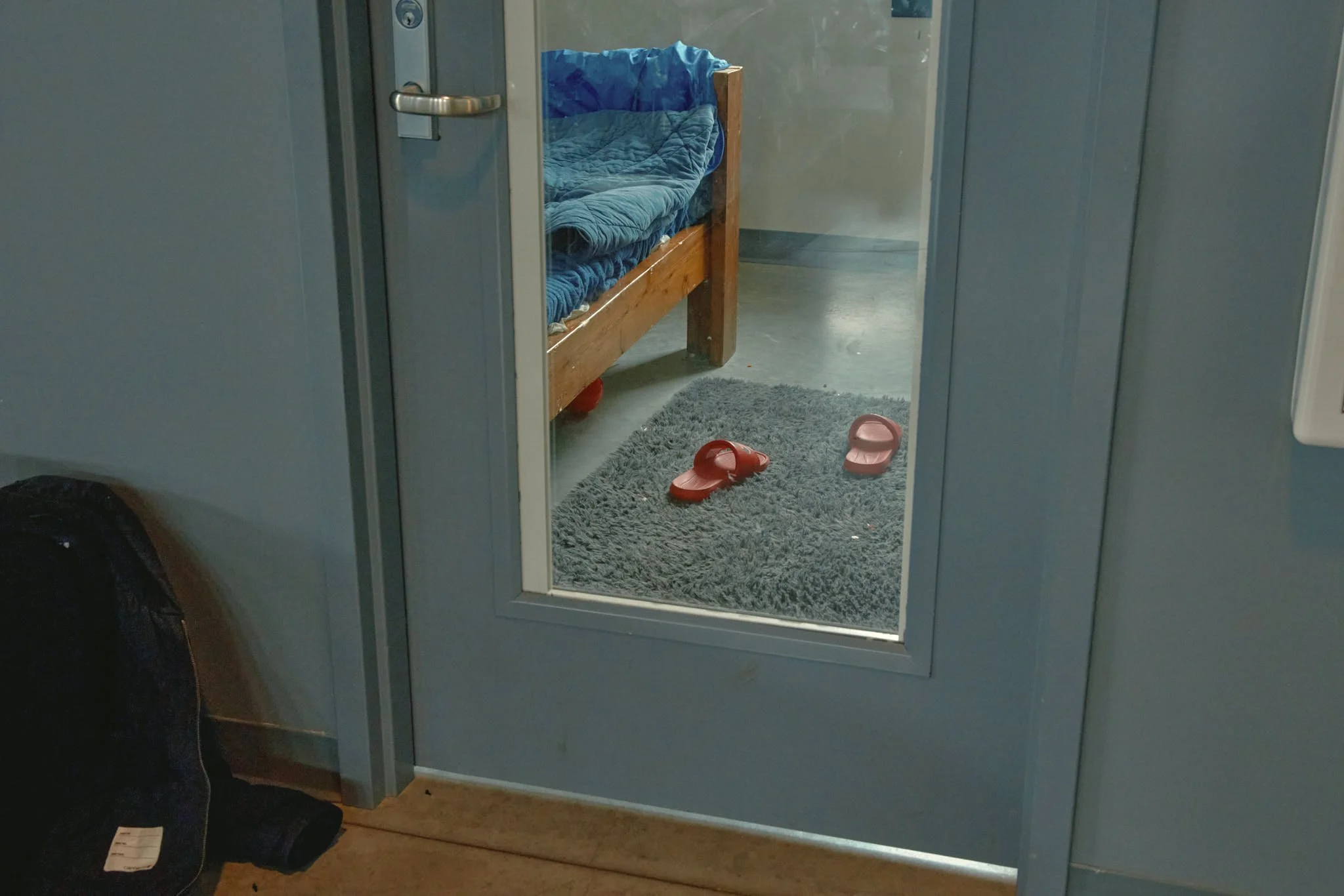 View through a doorway showing a small room with a wooden bed, a gray rug, and a pair of red sandals on the rug.