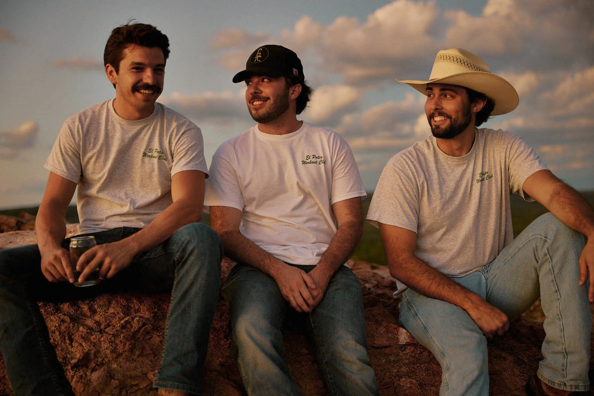 Three men sitting on a rock outdoors at sunset, smiling and talking, wearing casual clothes and the man on the right wearing a cowboy hat.
