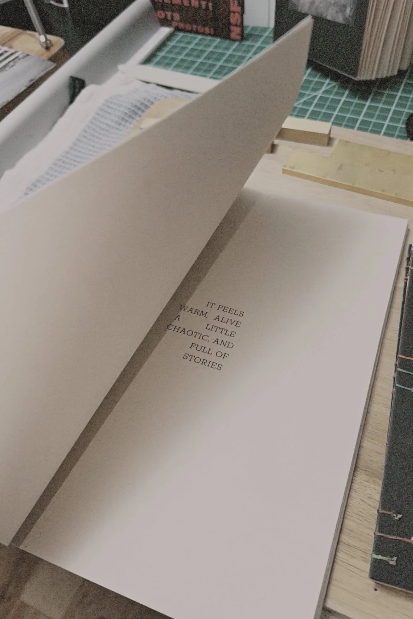 Open book with printed text on the right page, reading 'IT FEELS WARM, ALIVE A LITTLE CHAOTIC, AND FULL OF STORIES', on a wooden desk with other books and stationery.
