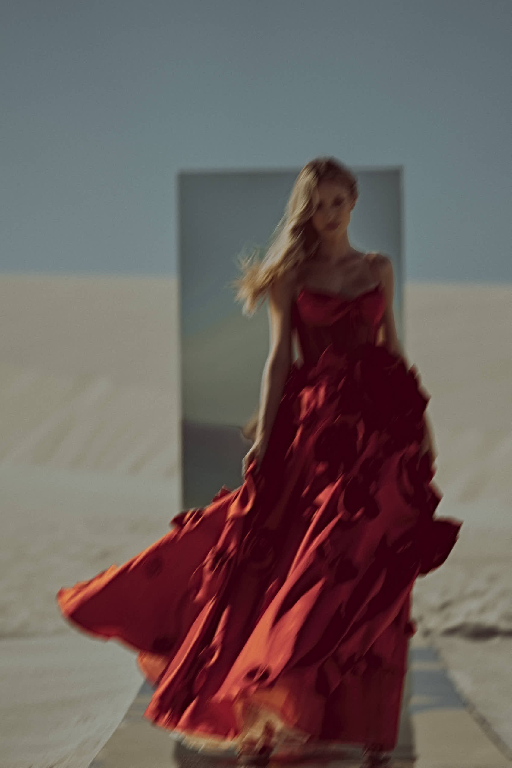 A woman in a flowing red dress walking in a sandy or beach area with a rectangular mirror in the background.