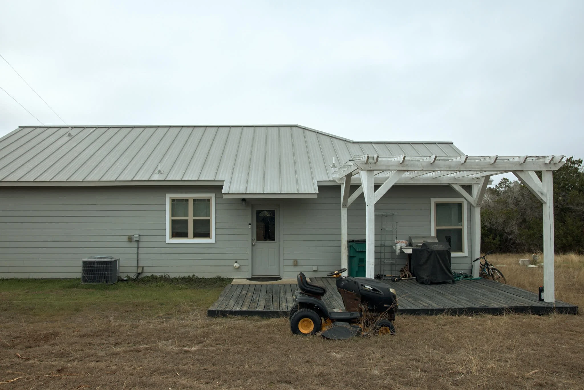 Back of a gray house with a metal roof, featuring a small porch with a wooden deck, a black riding mower, and outdoor items including bikes, a grill, and trash bins. The yard is dry with sparse grass and bushes in the background under an overcast sky.