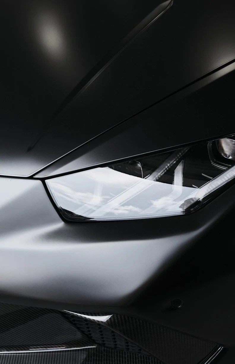 Close-up of a black luxury sports car's front, showing the sleek, angular headlight and matte black bodywork with hints of carbon fiber.