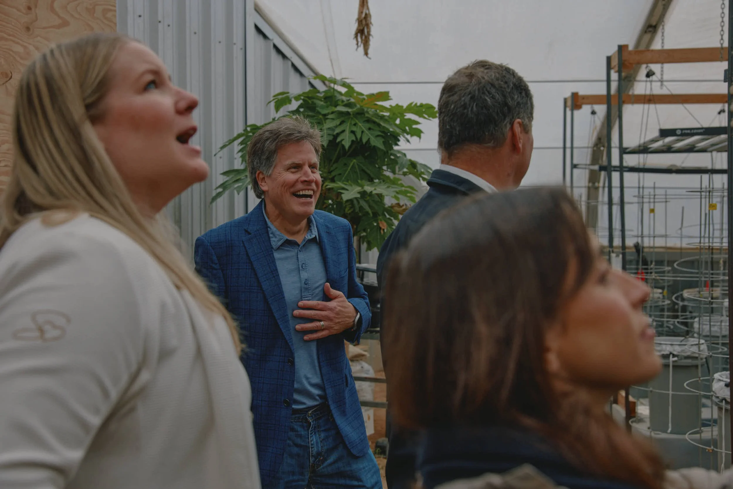 Group of people laughing and smiling indoors, near plants and greenhouse equipment.