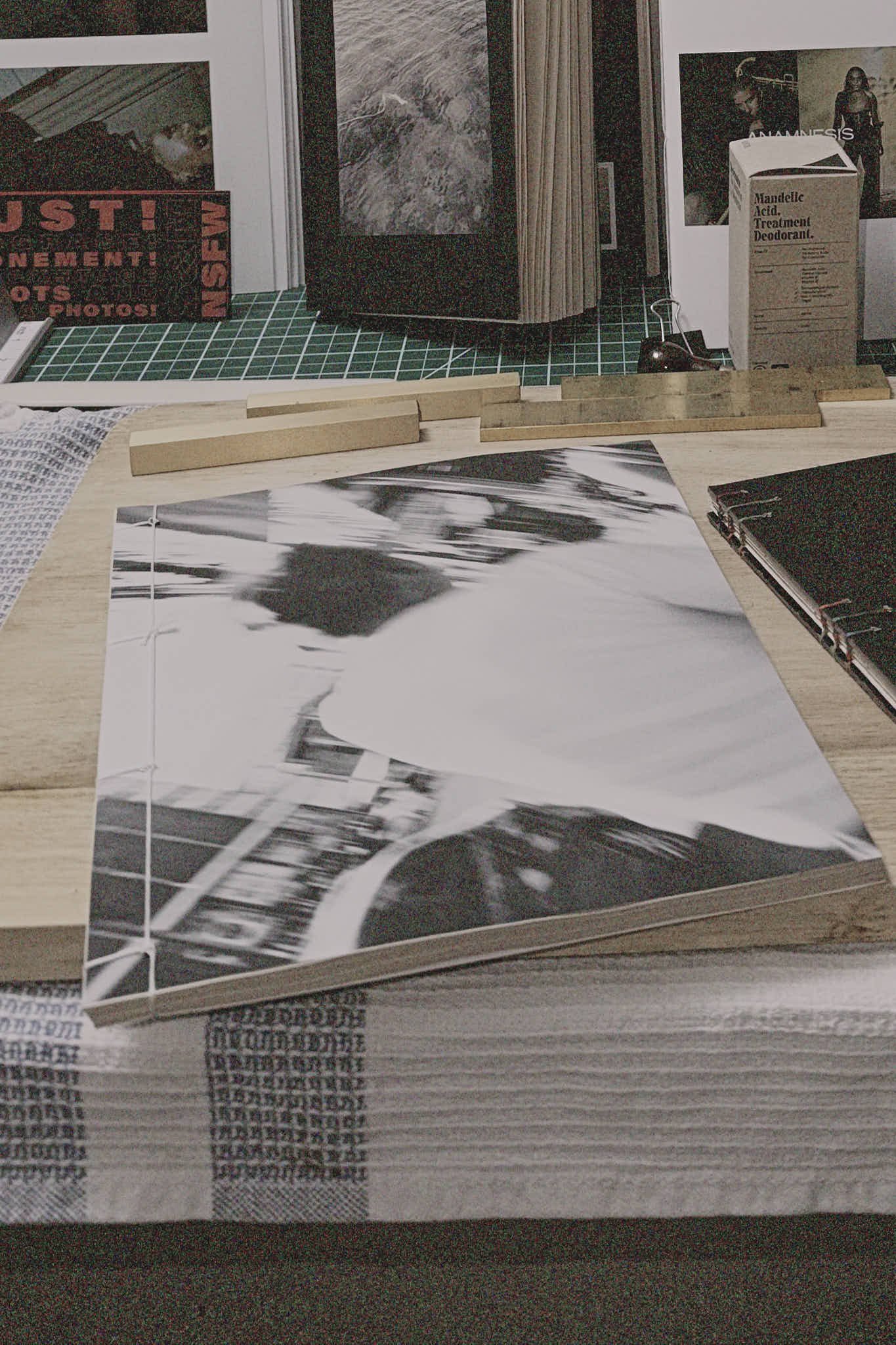 A black-and-white photograph of a person with glasses on a cluttered desk, surrounded by art supplies, photographs, and framed pictures.