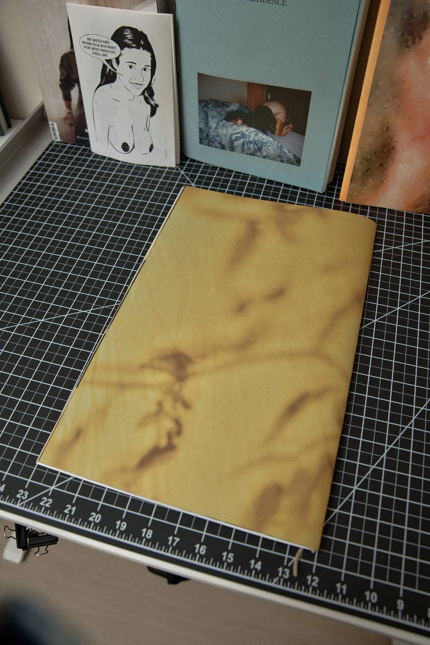 A black and white illustration of a woman with speech bubble, a photo of a person lying on a bed, and several art books on a black grid cutting mat.