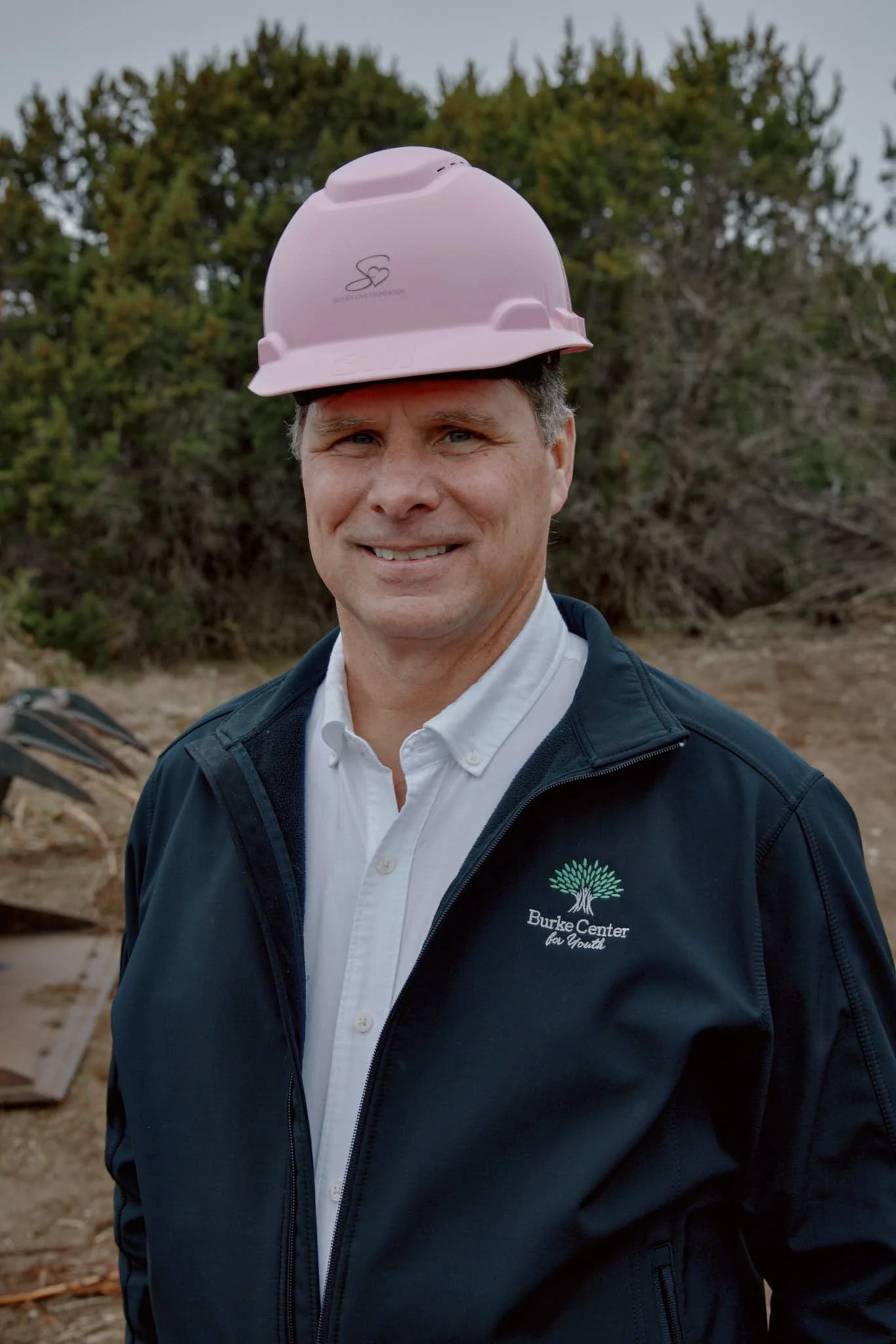 A man outdoors wearing a pink safety helmet and a navy jacket with the Burke Center for Youth logo, smiling at the camera.