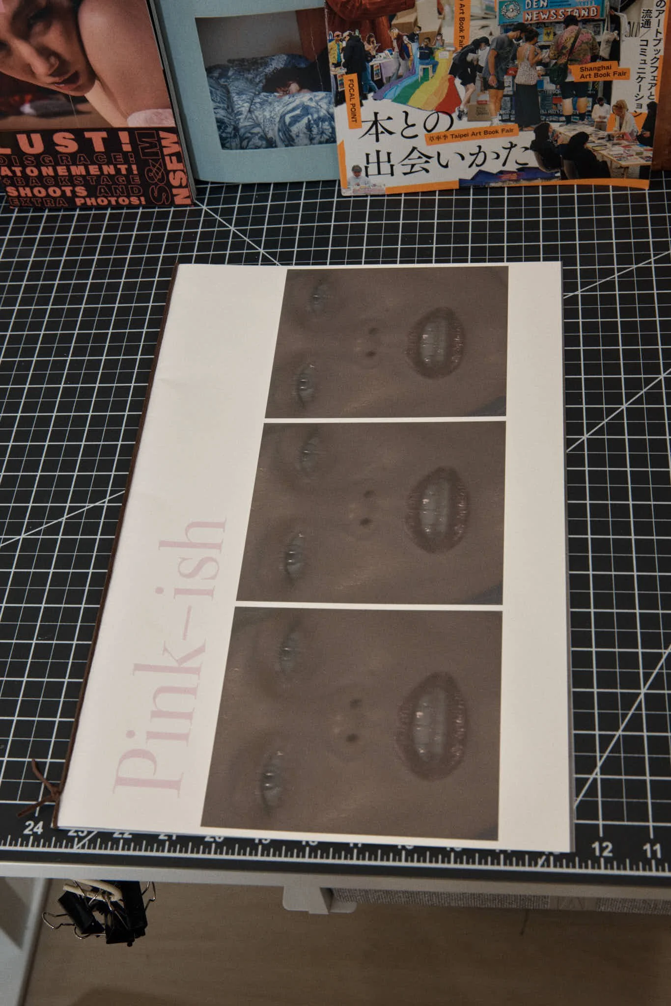 A brochure with three close-up photographs of a person's face focusing on the eyes and mouth, and the text 'Pink-ish' on the left side, lying on a black grid cutting mat.