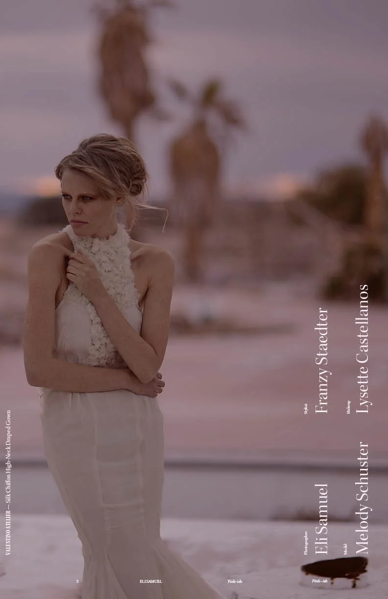 A woman standing on a sandy beach with palm trees in the background, wearing a white dress and holding her arm across her chest.