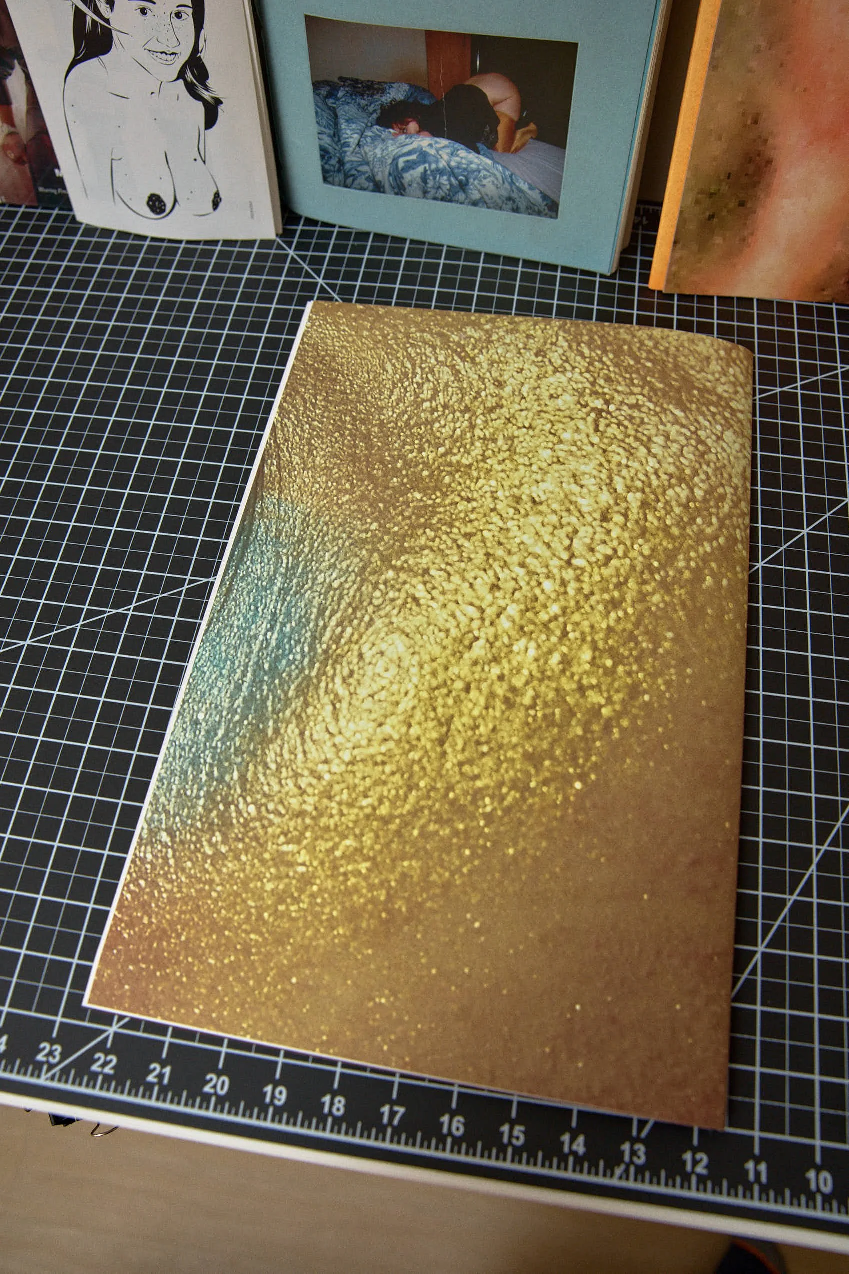 A gold-colored book cover with a textured, speckled pattern. In the background, there are framed photos and artwork, including a sketch of a woman and a photo of a person sleeping on a bed.