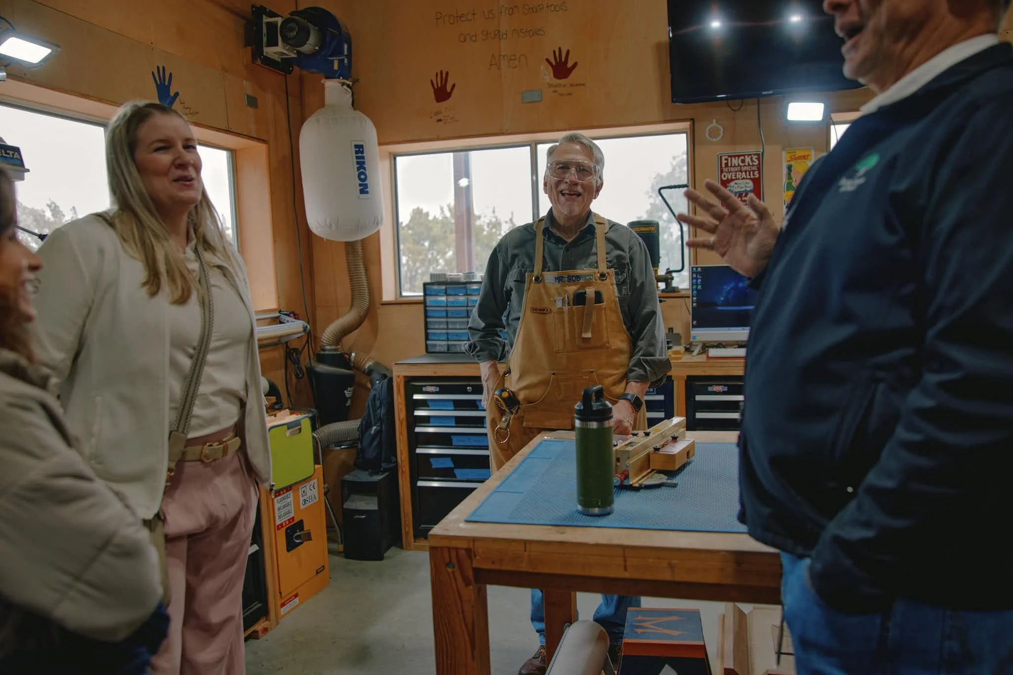 Group of people talking and smiling in a woodworking shop. One man in a tan apron is standing behind a workbench, while others are gathered around, engaged in conversation.
