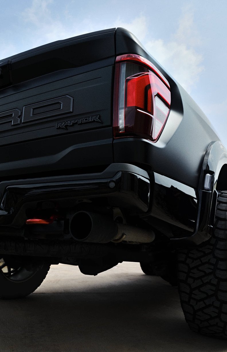 Close-up of the rear end of a black Ford Raptor pickup truck, showing the tail light, tire, and exhaust pipe against a cloudy sky.