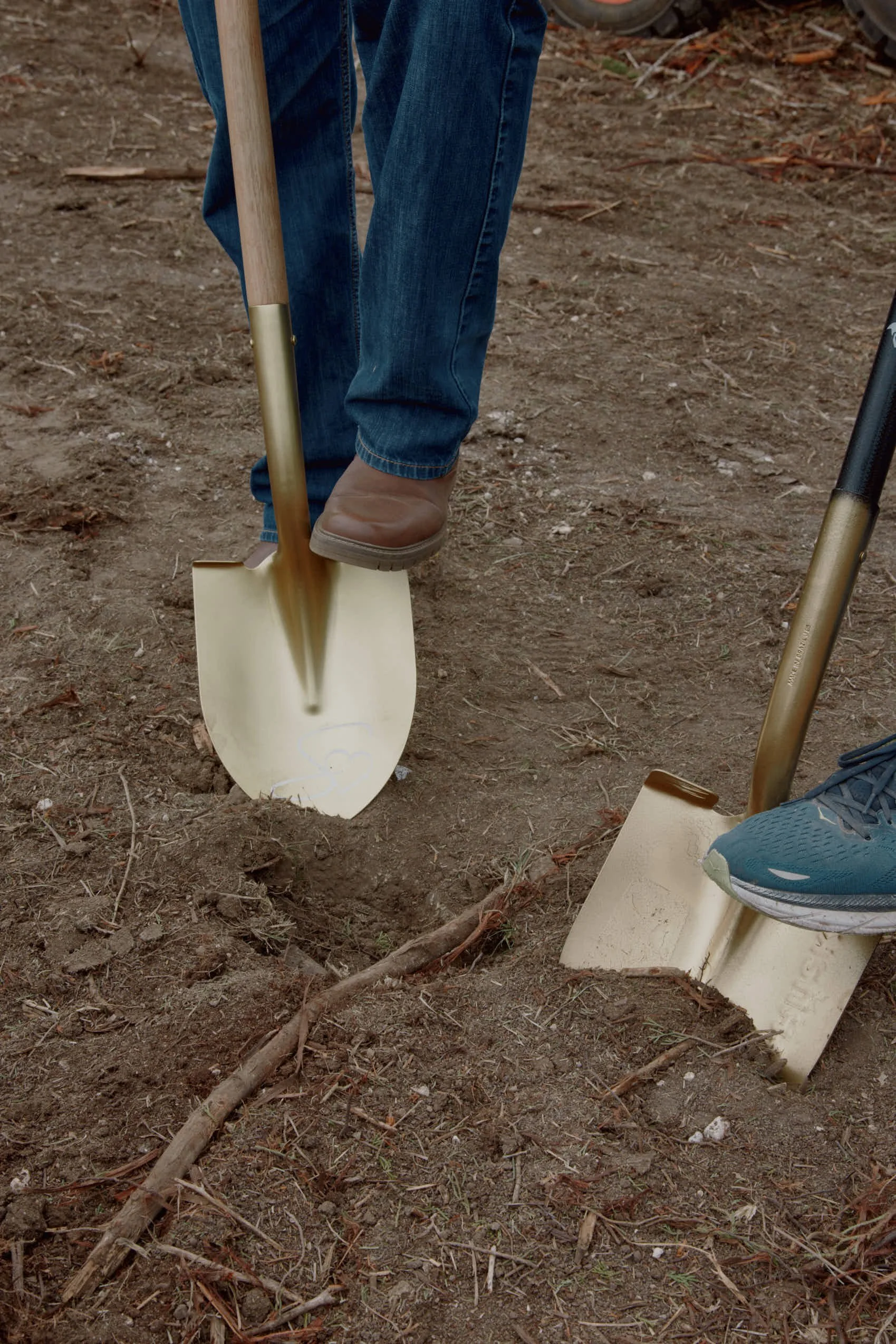 Close-up of two people digging into the dirt with small shovels, one wearing brown boots and blue jeans, the other wearing a blue shoe, on an outdoor dirt surface.
