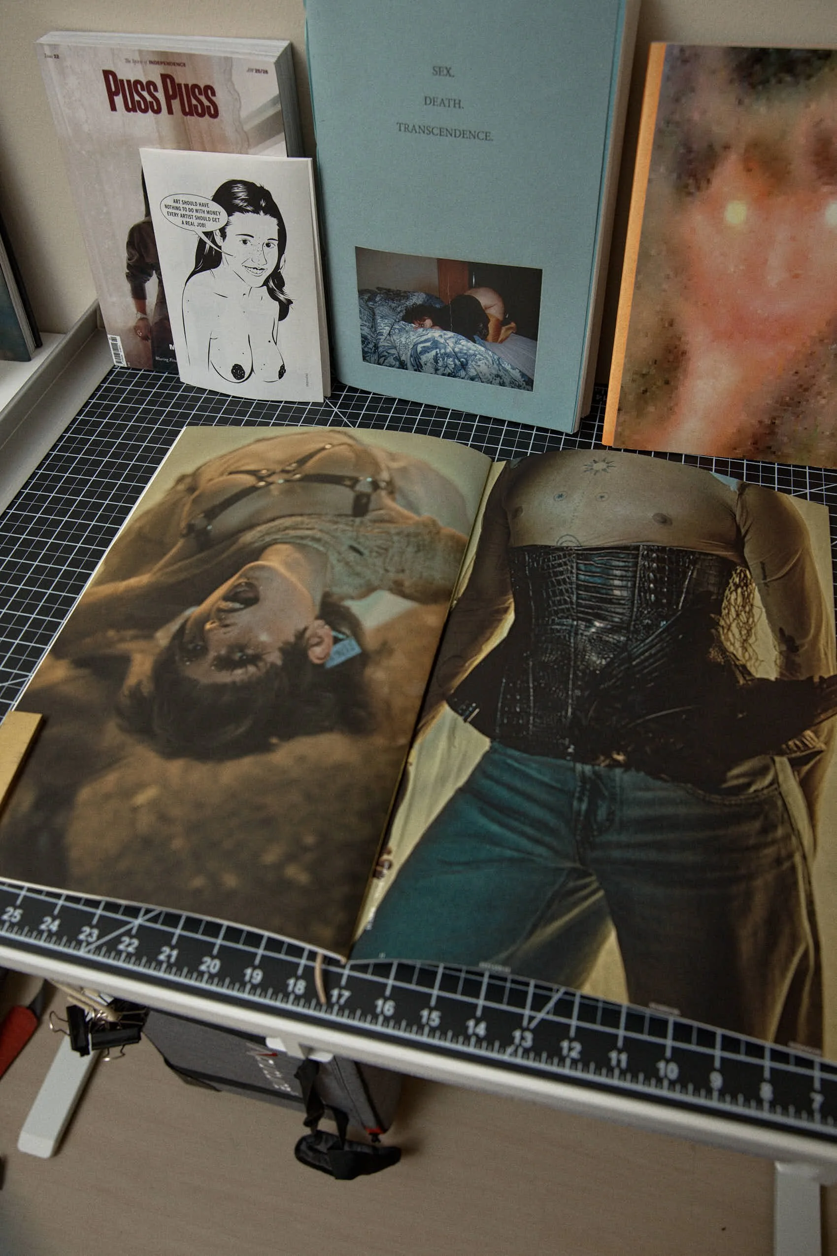 Open magazine with provocative imagery of women, accompanied by other magazines and artwork displayed on a table.