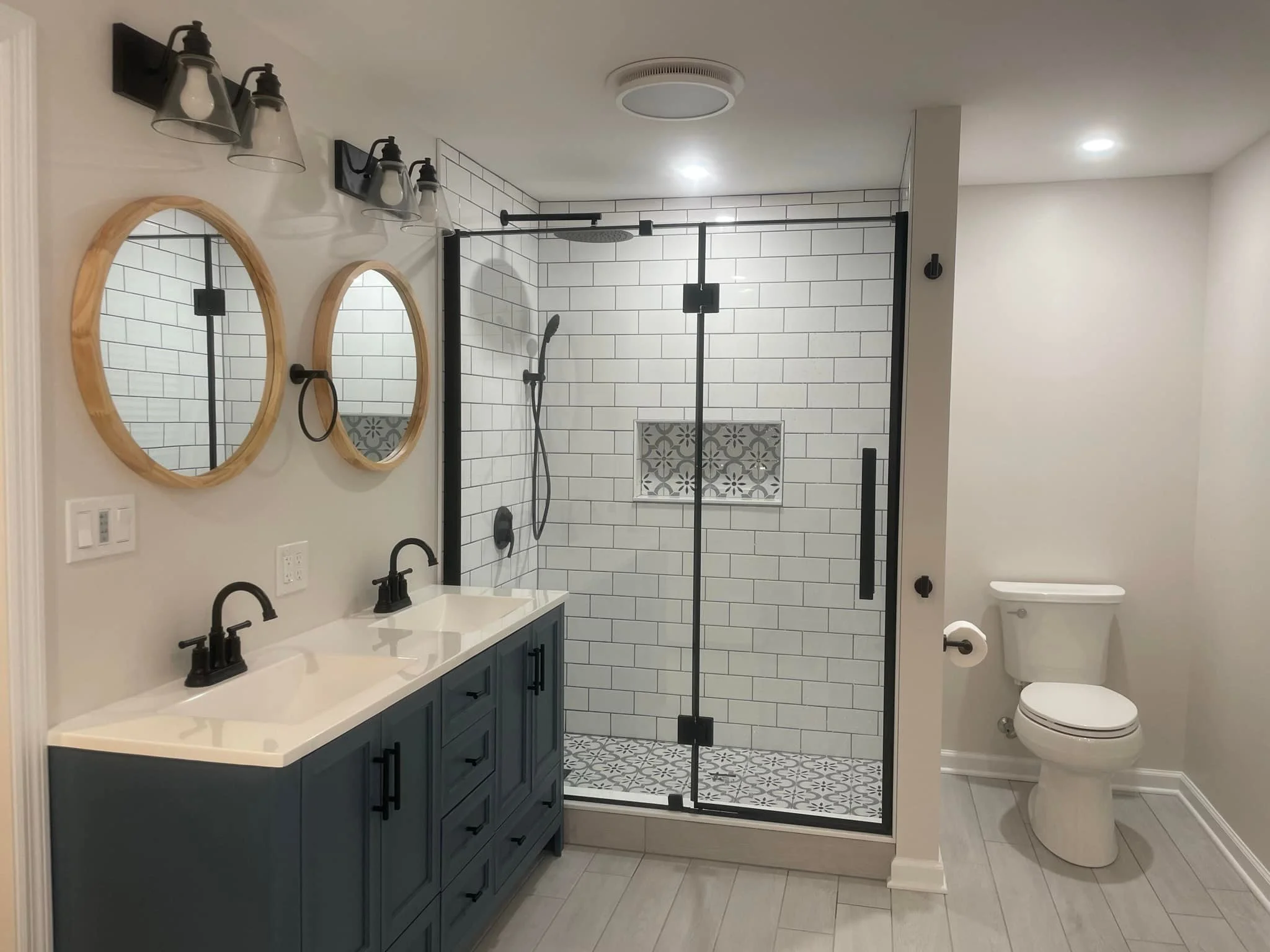 Modern bathroom with black and white hexagonal tile floor, wood vanity with black countertop and gold fixtures, oval mirror, wall light with two round bulbs, toilet, and walk-in shower with green tiled wall, built-in shelf, small window, and black shower fixtures.