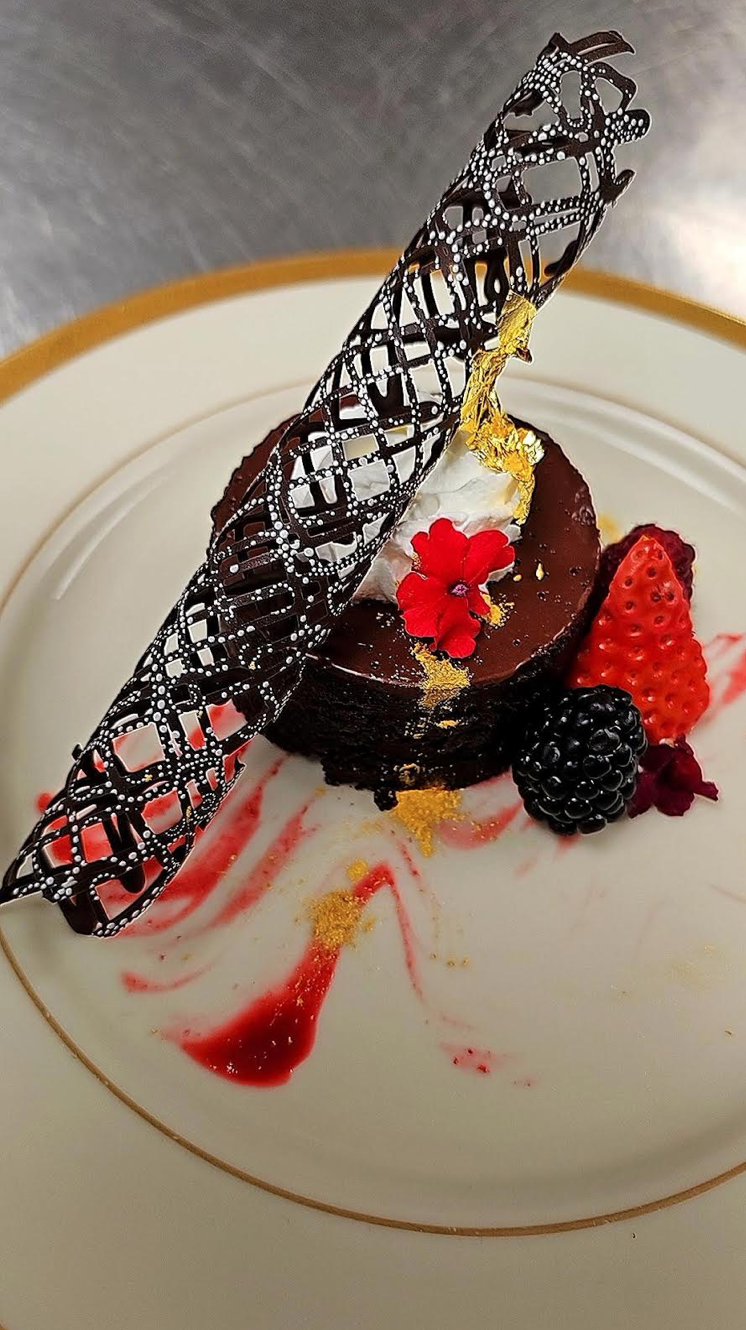 A gourmet chocolate dessert garnished with fresh berries, edible gold leaf, whipped cream, a decorative chocolate lattice, and a red flower, served on a white plate with red sauce and gold accents.